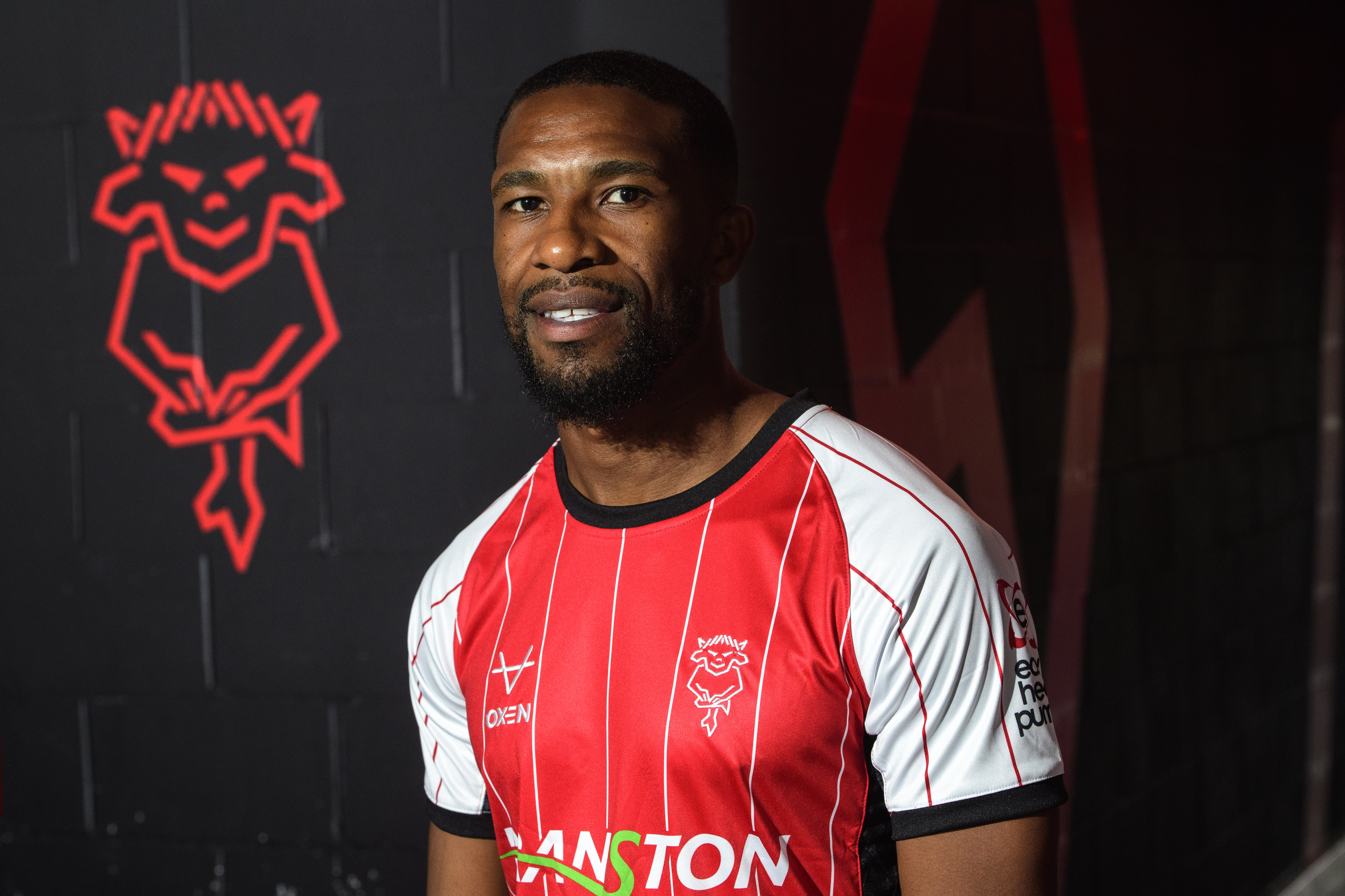 Tendayi Darikwa poses in the tunnel at the LNER Stadium. He is wearing a red home shirt and in the background the the Lincoln City badge.