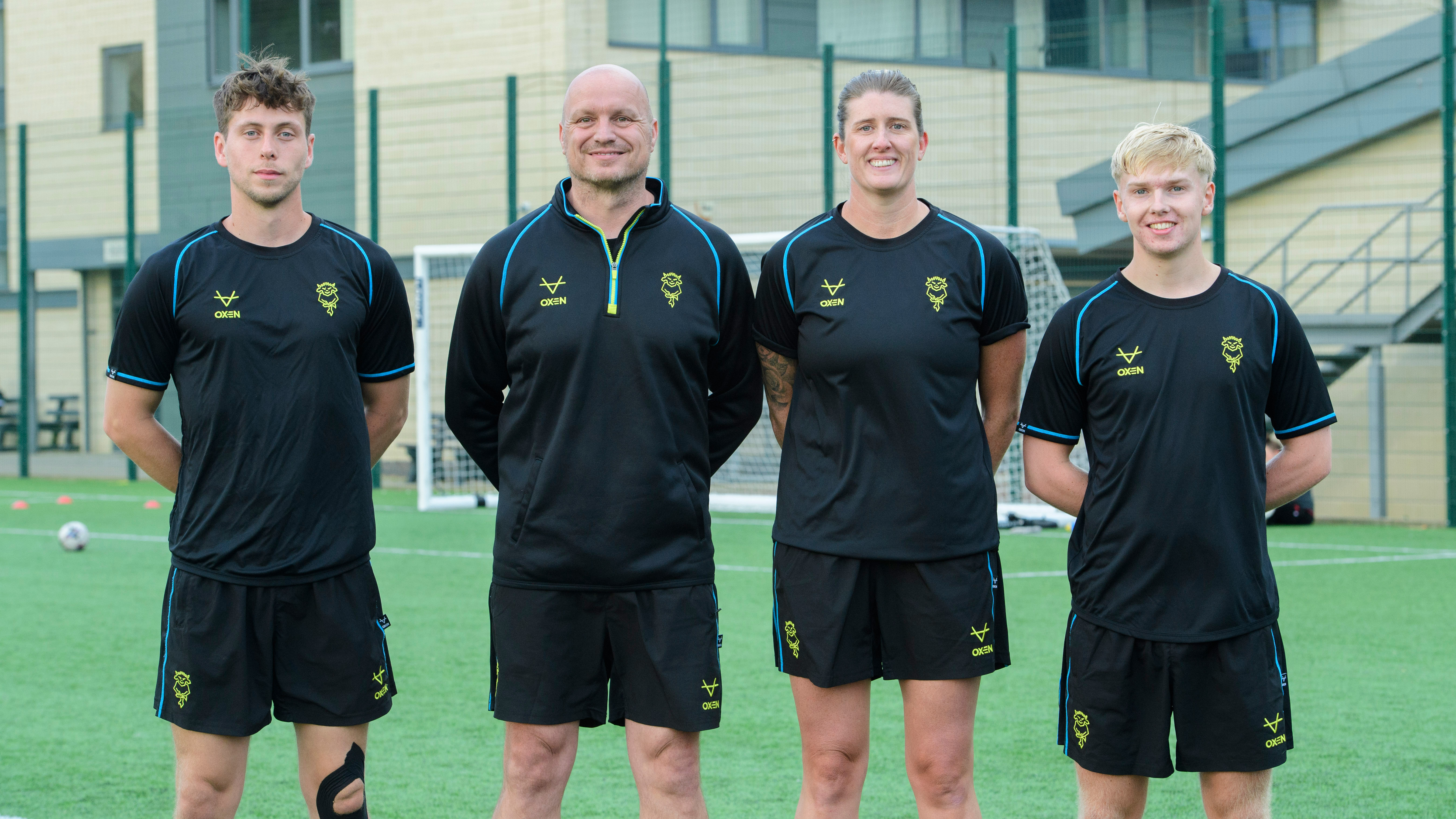 Four members of Lincoln City Women coach staff pose in front of a football pitch. They are all wearing black training kit. 