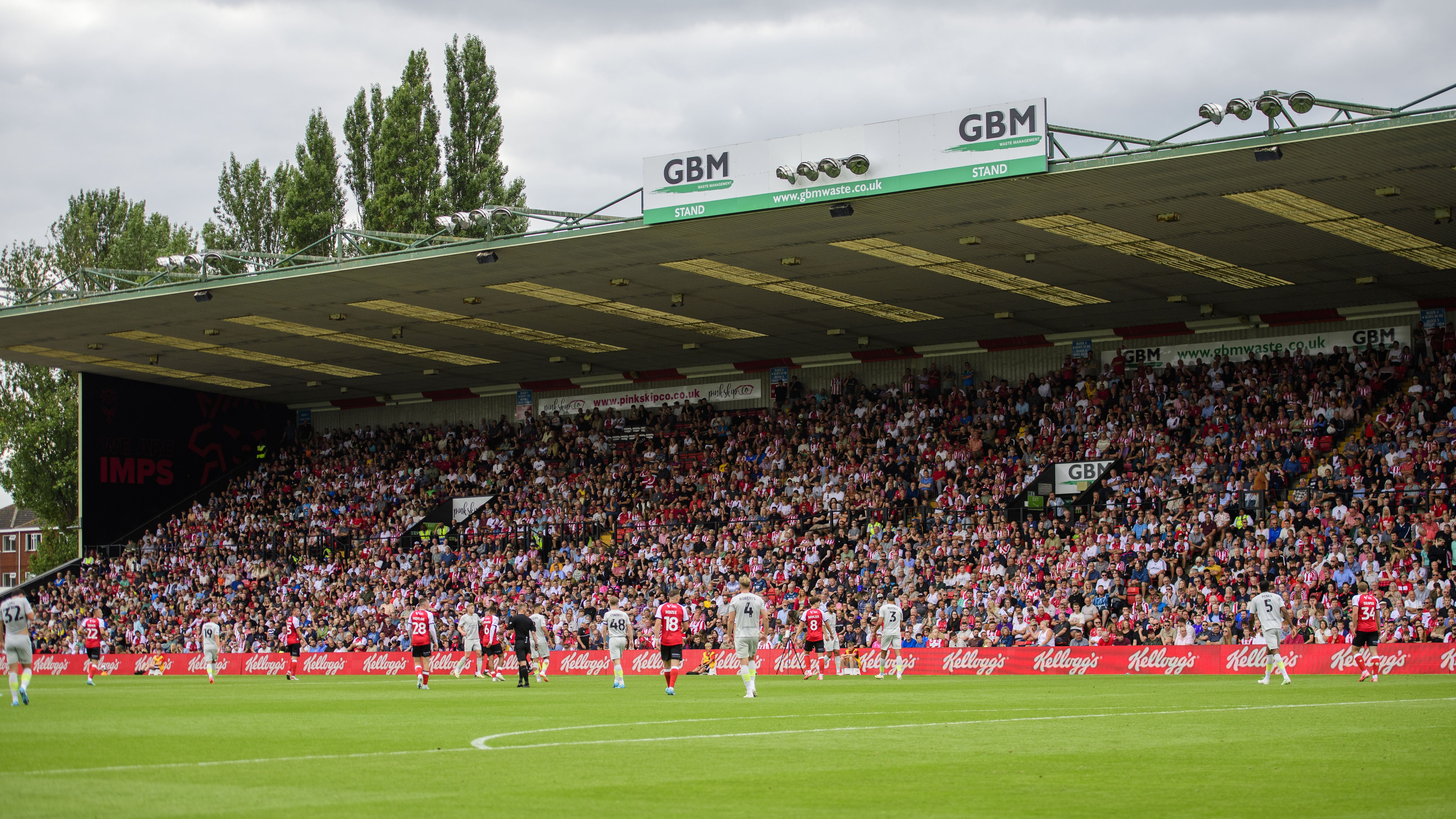 Lincoln City supporters in the GBM Stand. In the foreground there are several players both of Lincoln City, wearing red, and an opposition team wearing white.