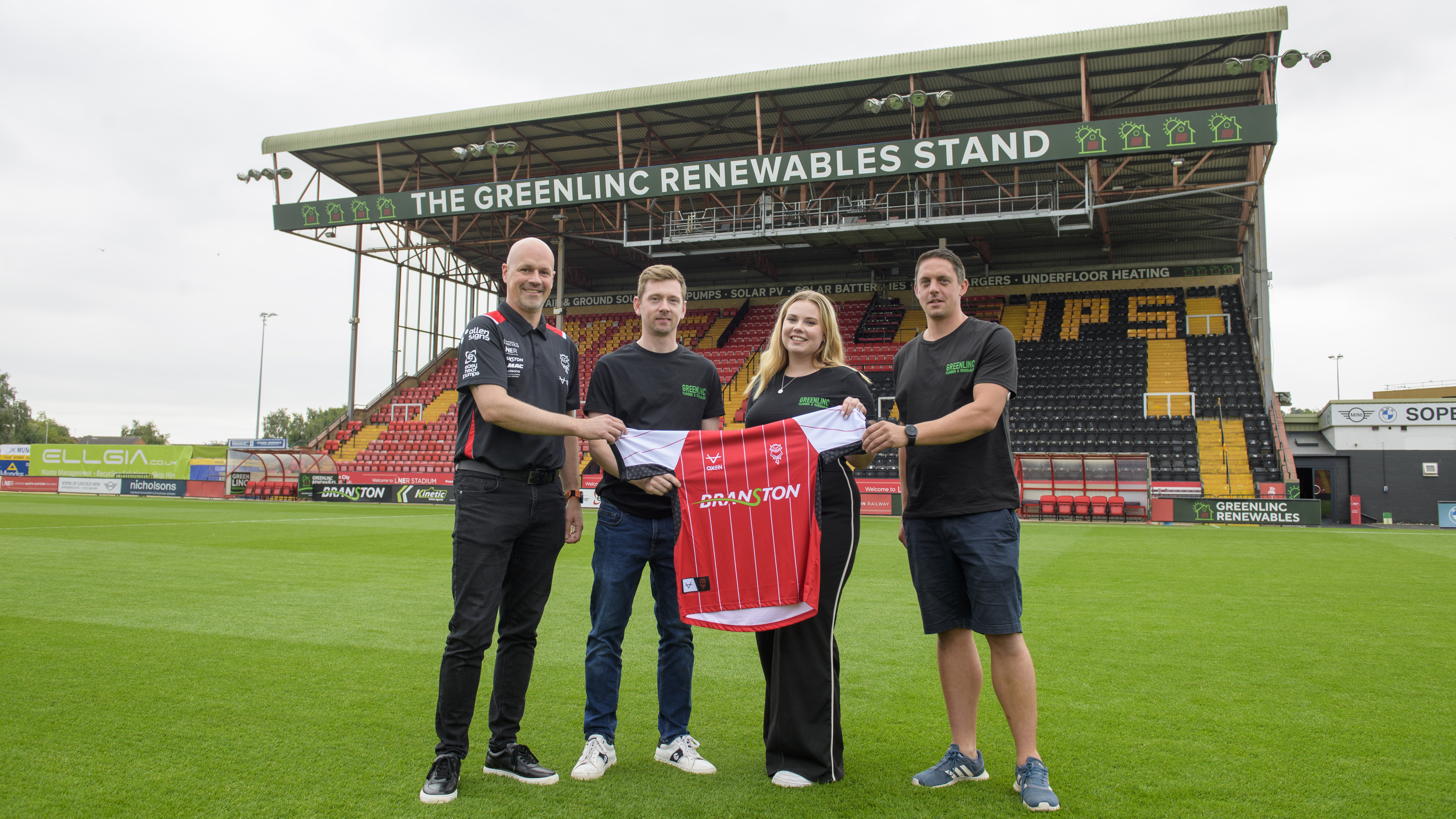 Four people stand on a football pitch holding a red Lincoln City shirt. Behind them is a stand which has a sign reading "Greenlinc Renewables Stand"