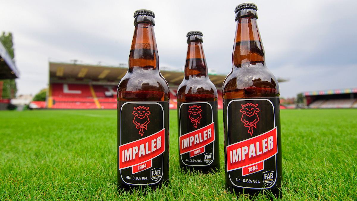 Three bottles of Impaler 1884 beer on the green grass of the pitch at the LNER Stadium. The GMB Stand can seen in the background.