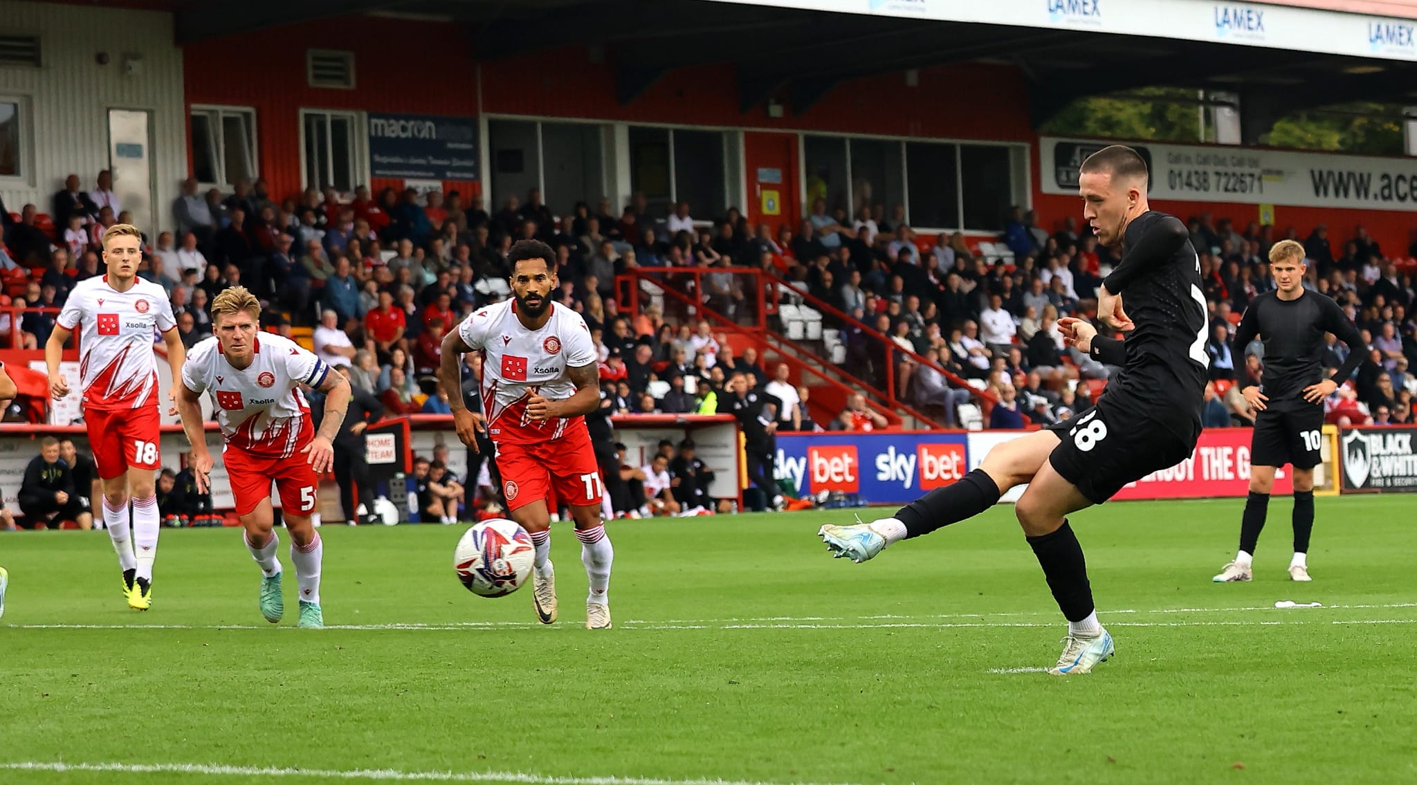 Jack moylan, wearing an all black kit, kicks the ball with his right foot to score a penalty against Stevenage.