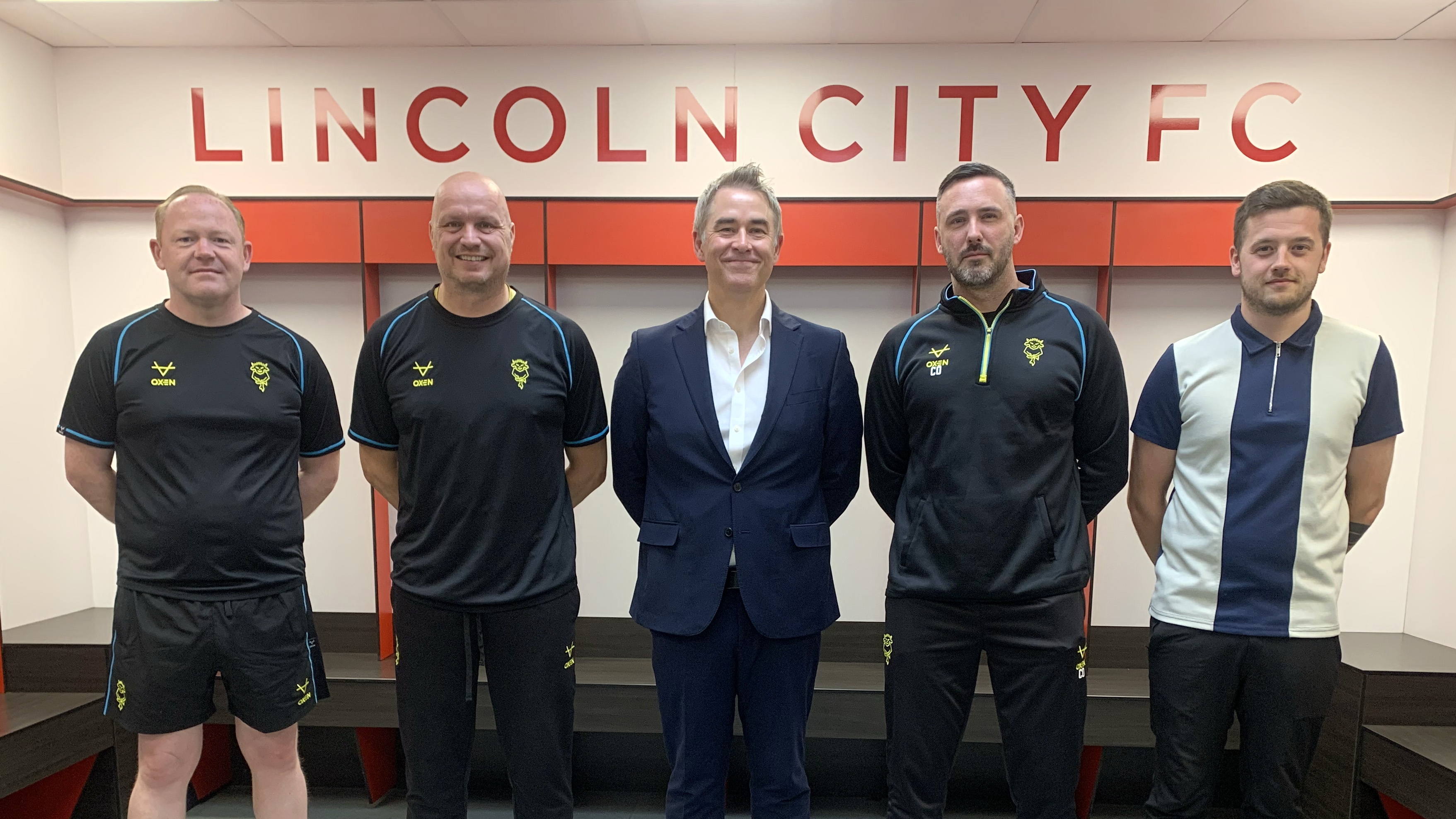 Marc Tracy, Jon Pepper, Jason Futers, Calum Oakenfold and Matt Murgett post in the changing room at the LNER Stadium. Above them is a sign which reads Lincoln City FC