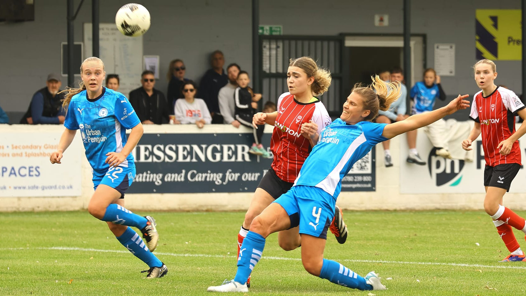 A Lincoln City Women player, wearing red, tries to control the football. There are Peterborough players, both wearing blue, on either side of her.