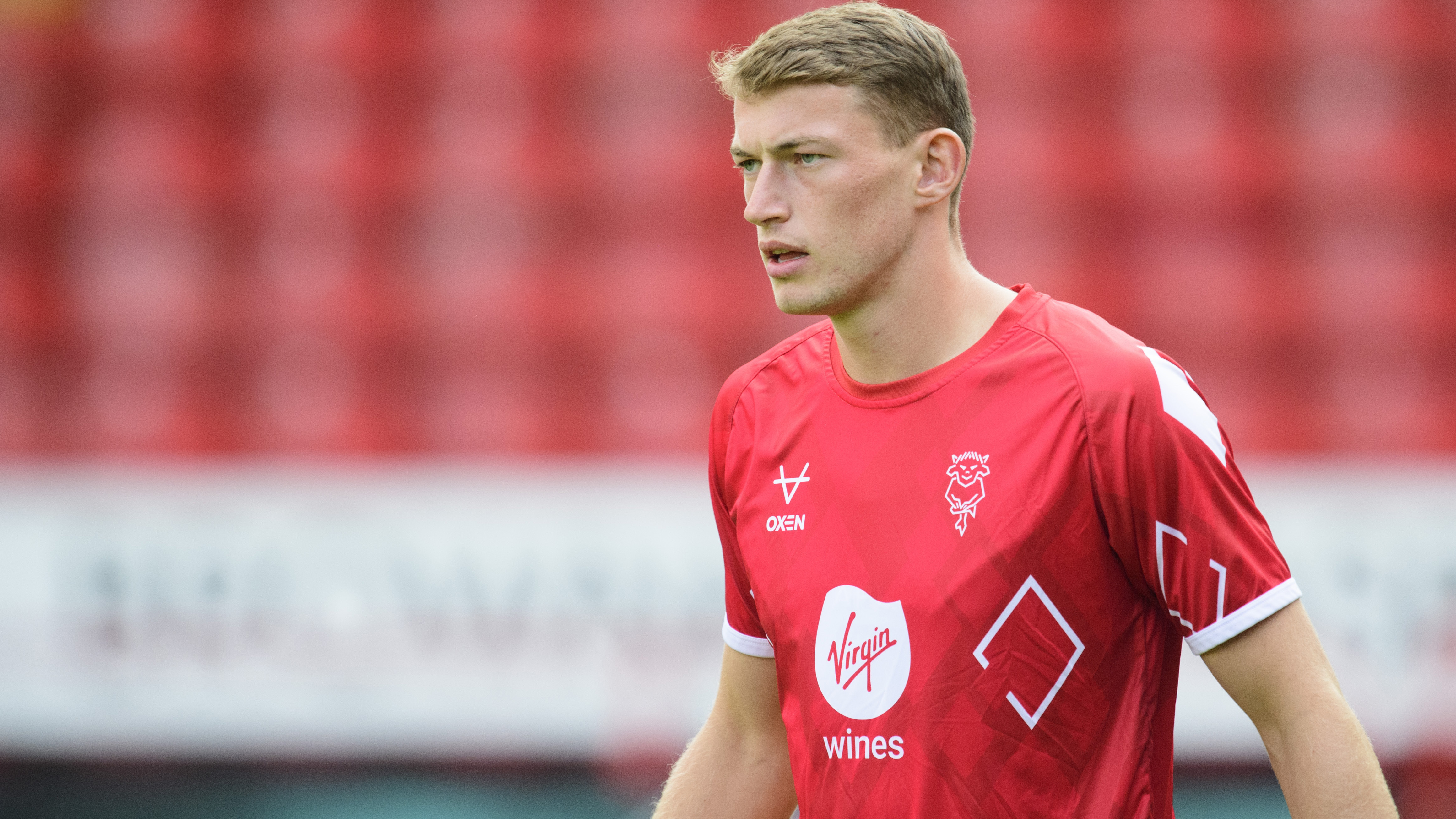 Zak Bradshaw is pictured wearing a red warm-up t-shirt. He's looking from the right of the picture into the middle. There are red seats in the football stand in the background.