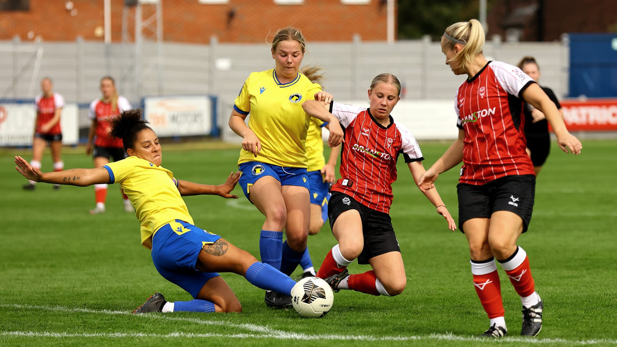 A Lincoln City Women player wearing a red top challenges for the ball with her right foot. She is competing with two Solihull Moors players who are wearing yellow tops