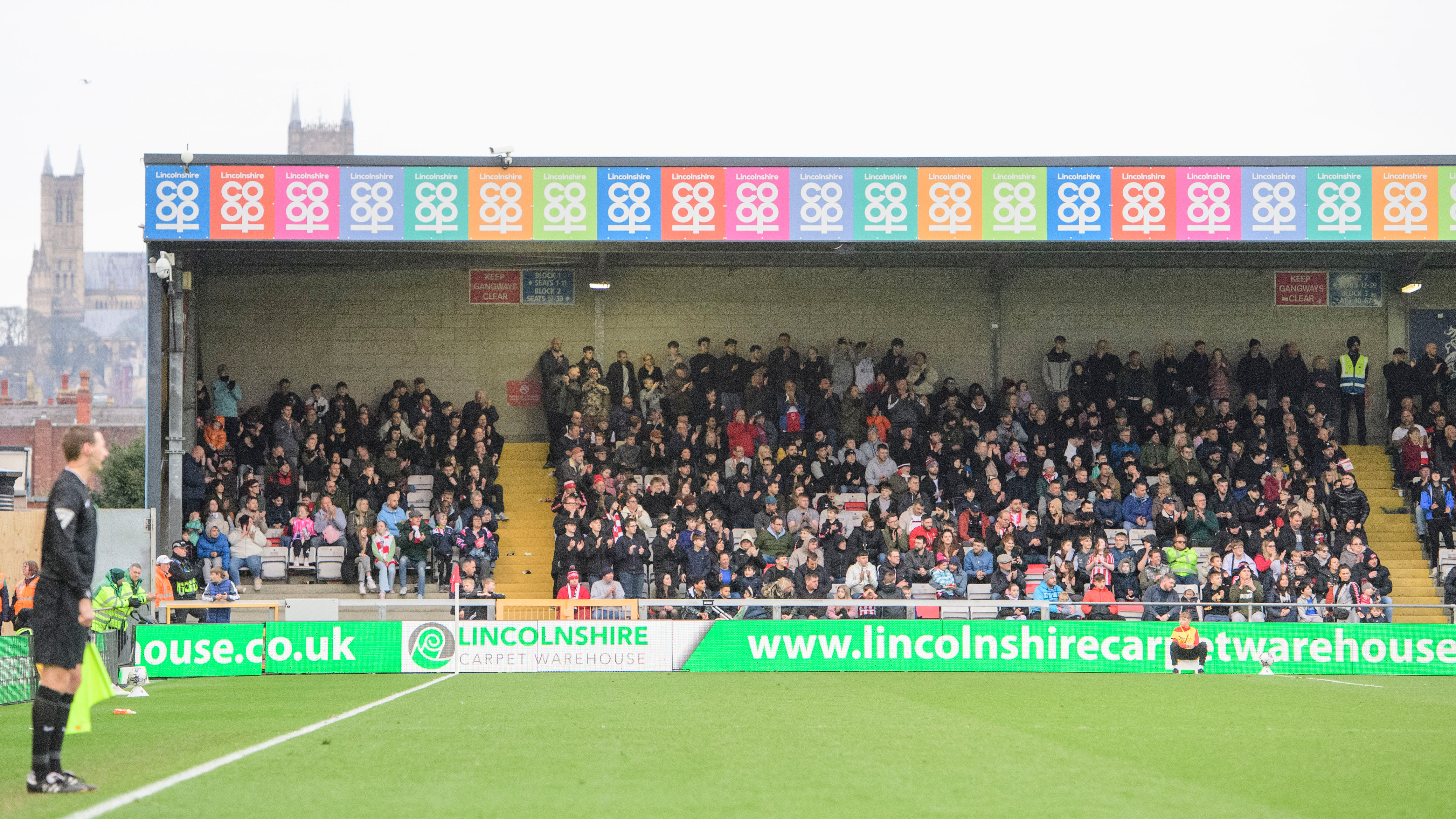 Supporters watch a game from the Stacey West Stand at the LNER Stadium. In the foreground is the pitch, and behind the stand you can see Lincoln Cathedral.