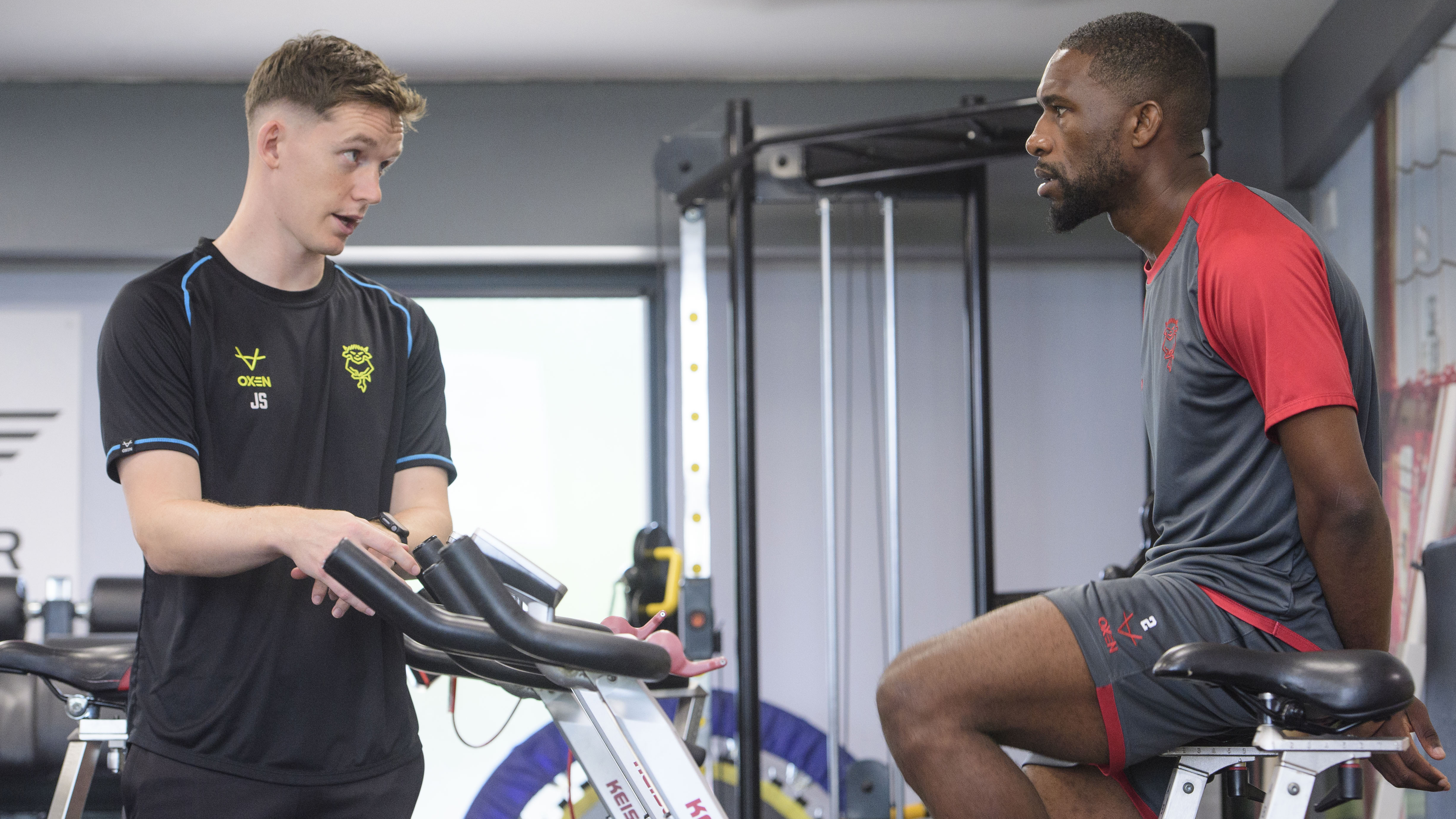 Two men in a gym. One man sits on an exercise bike talking to a trainer.