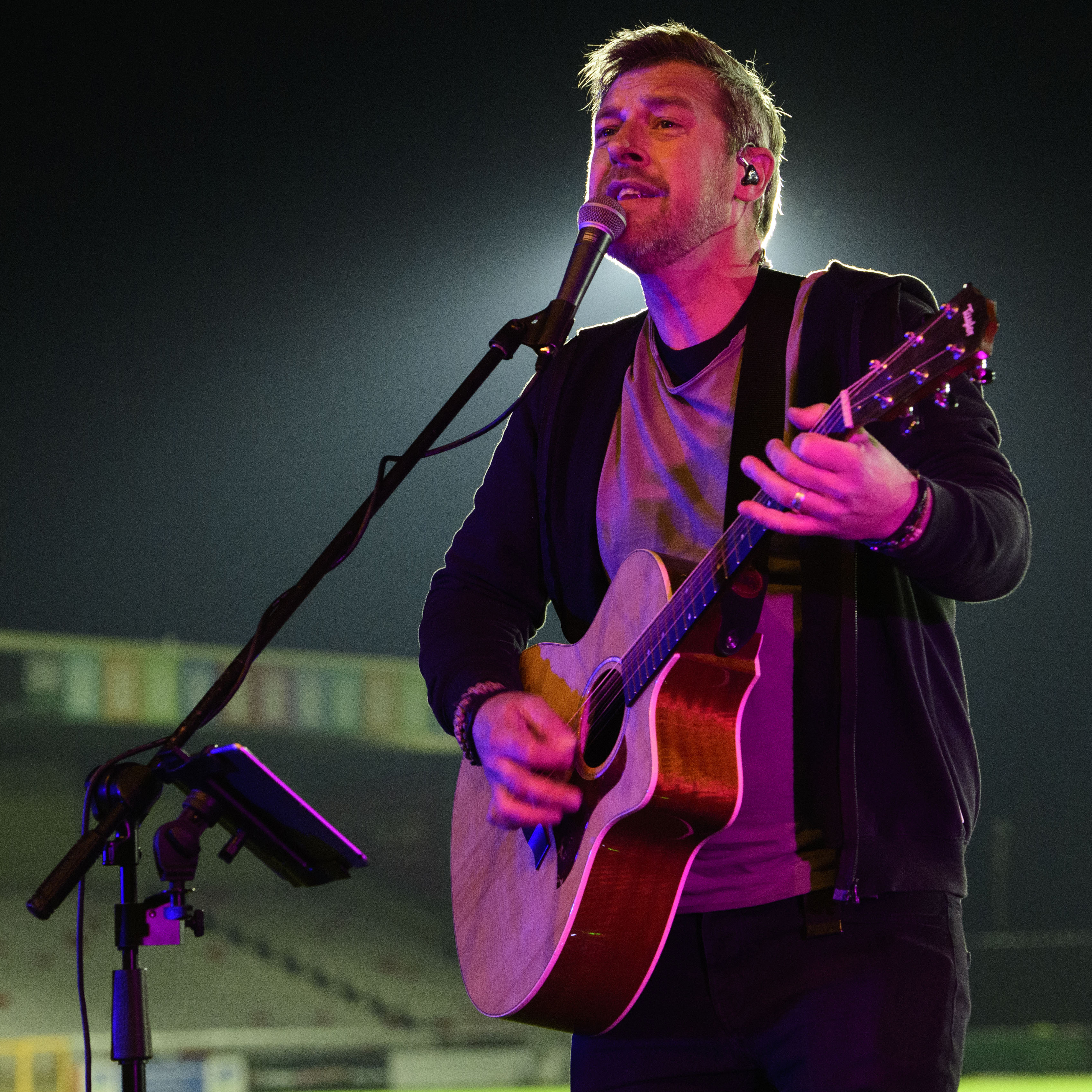 Graham Lindley performs an acoustic set. He is holding a guitar and singing into a microphone against a dark backdrop.