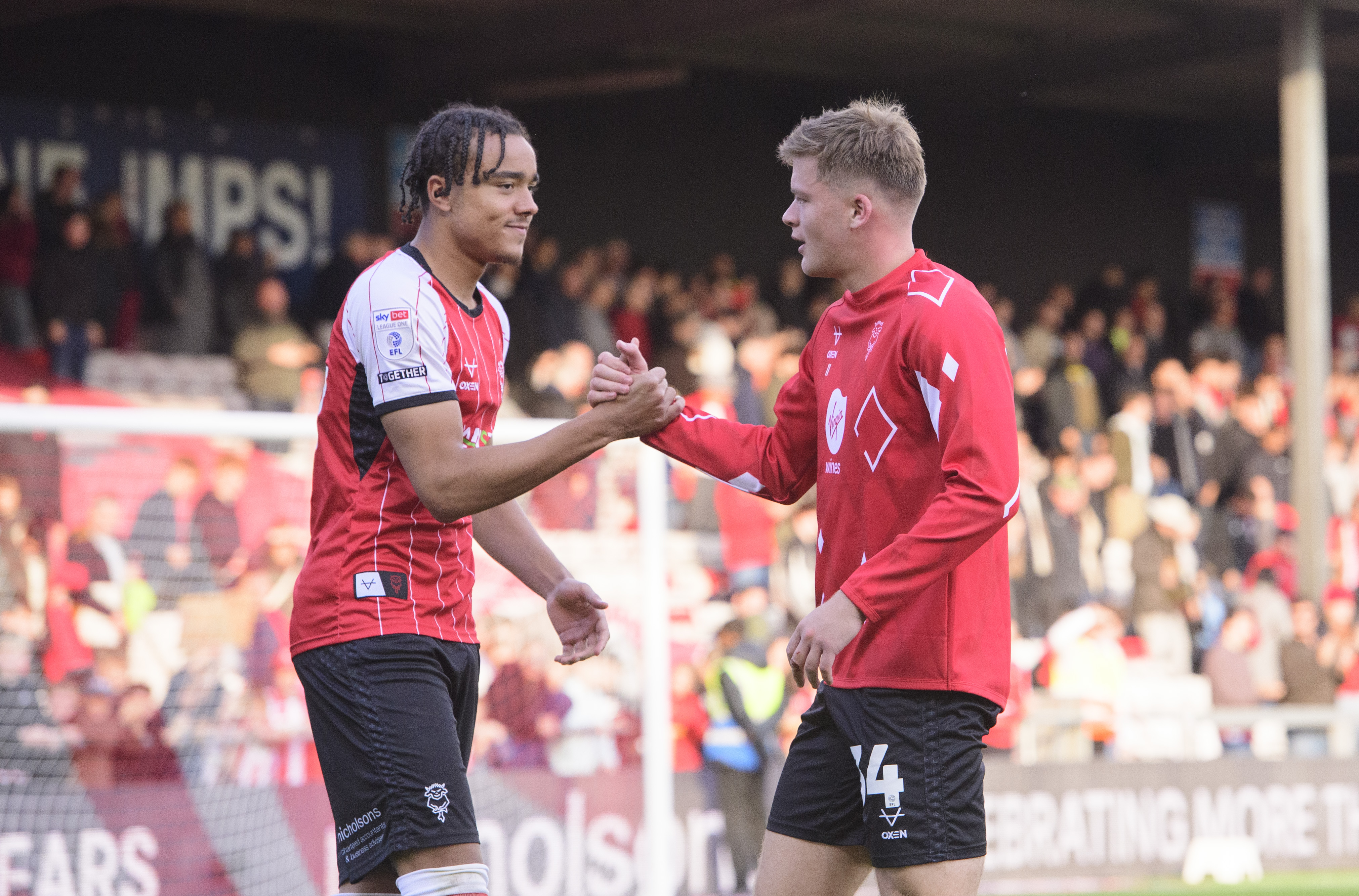 Jovon Makama and Freddie Draper clasp hands after celebrating Lincoln City's win over Leyton Orient. They are looking from the right of the image and both wearing red tops and black shorts.