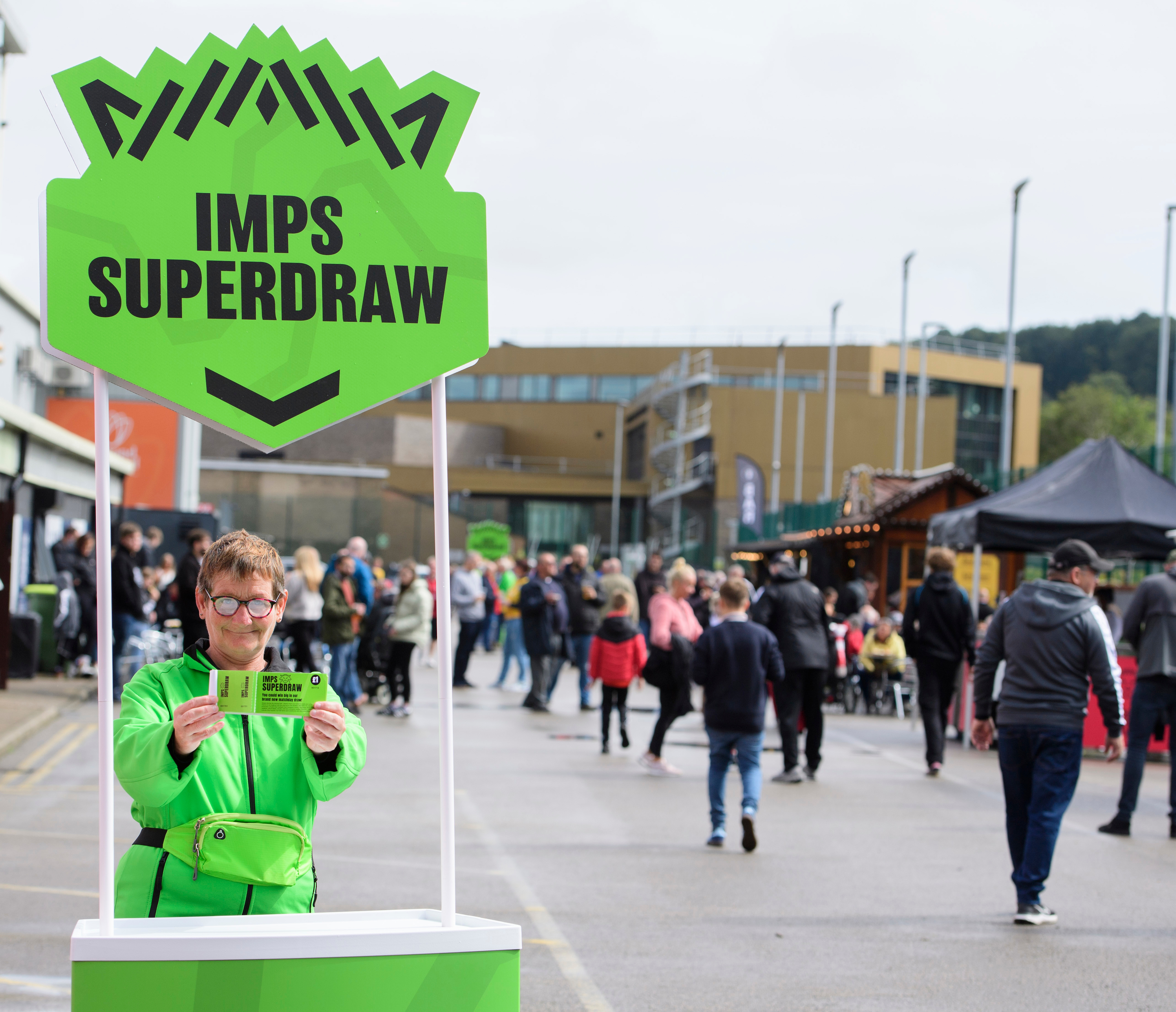 A person wearing bright green jacket holds up an Imps Superdraw ticket. They are stood behind a stand which is bright green and also reads "Imps Superdraw". People walk past in the background.