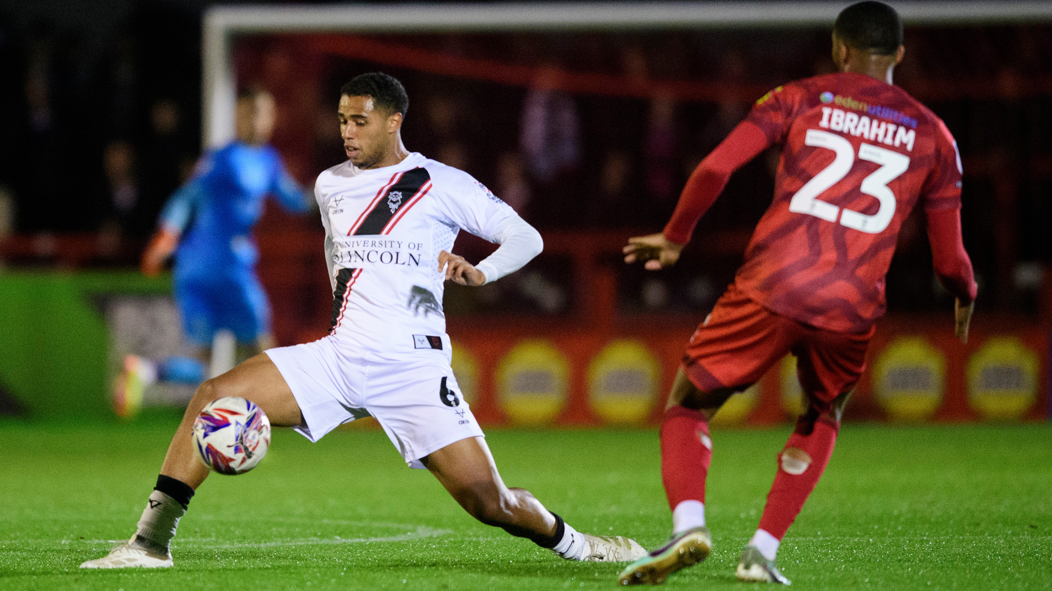 A footballer wearing an all-white kit intercepts the ball under pressure from another football in an all-red strip.