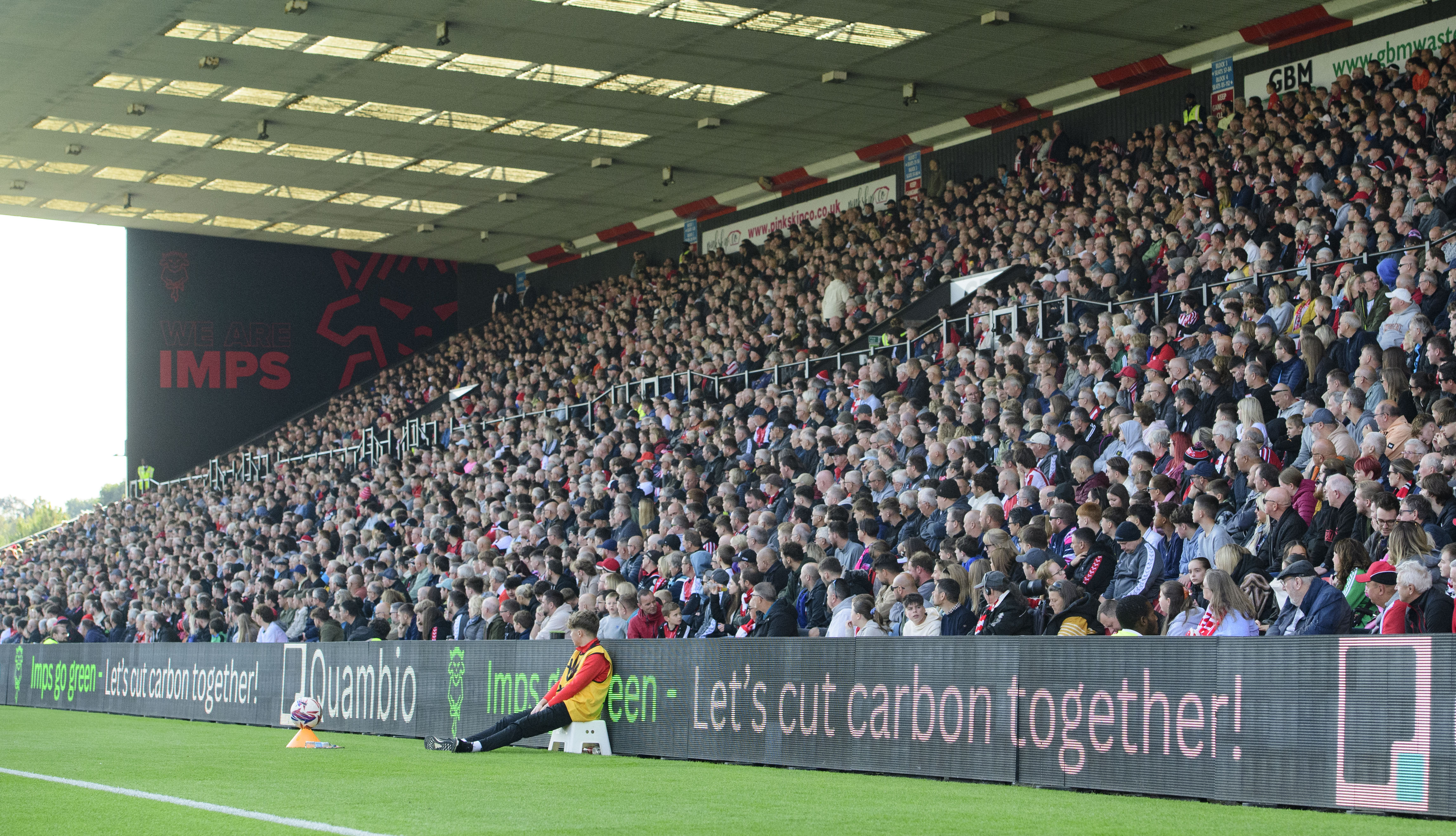 Lincoln City fans watch a game from a packed GBM Stand. At the back of the stand is a mural which reads "We Are Imps".