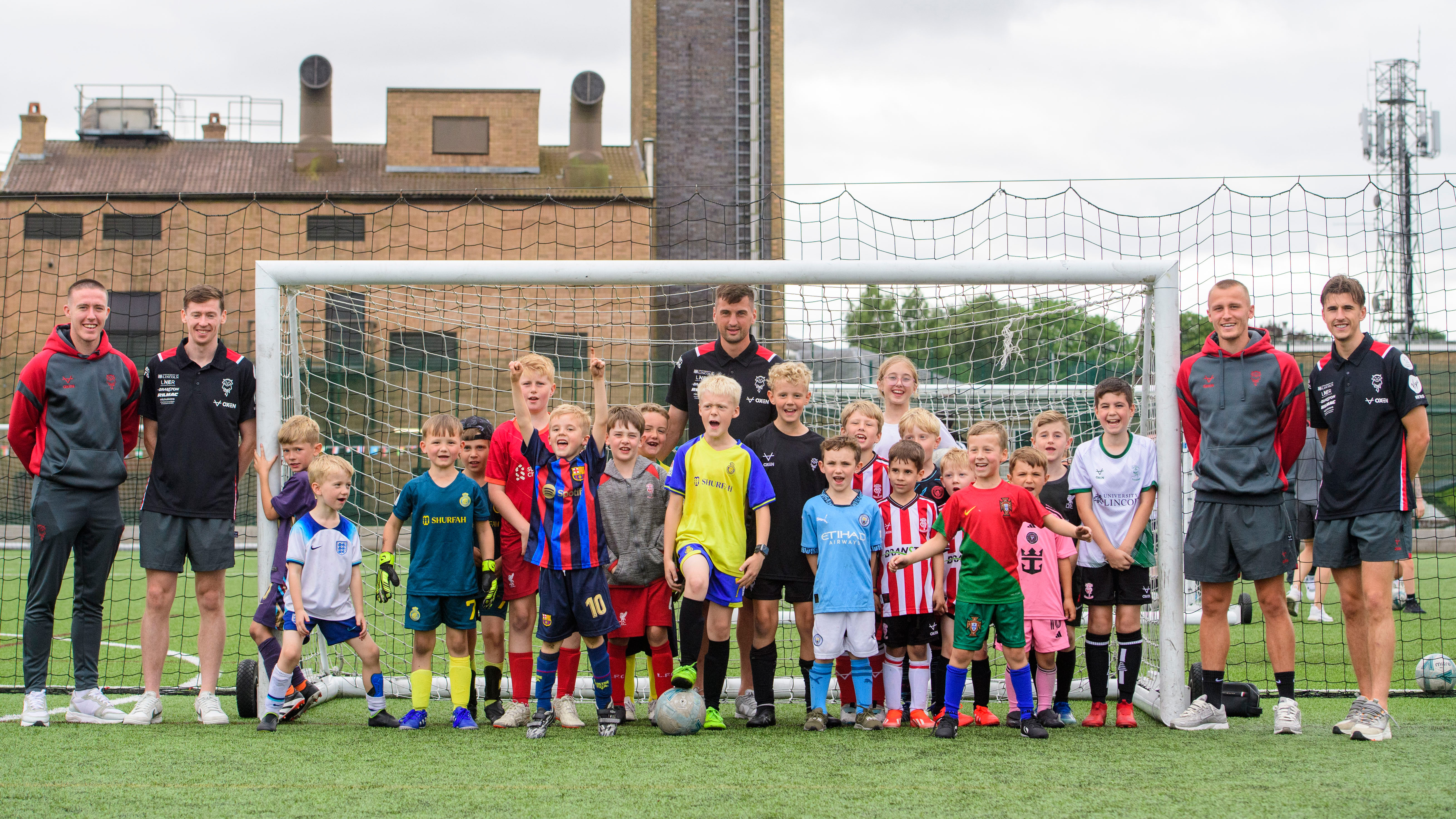 A group of around 20 children pose with five Lincoln City players. They are stood in front of a goal.