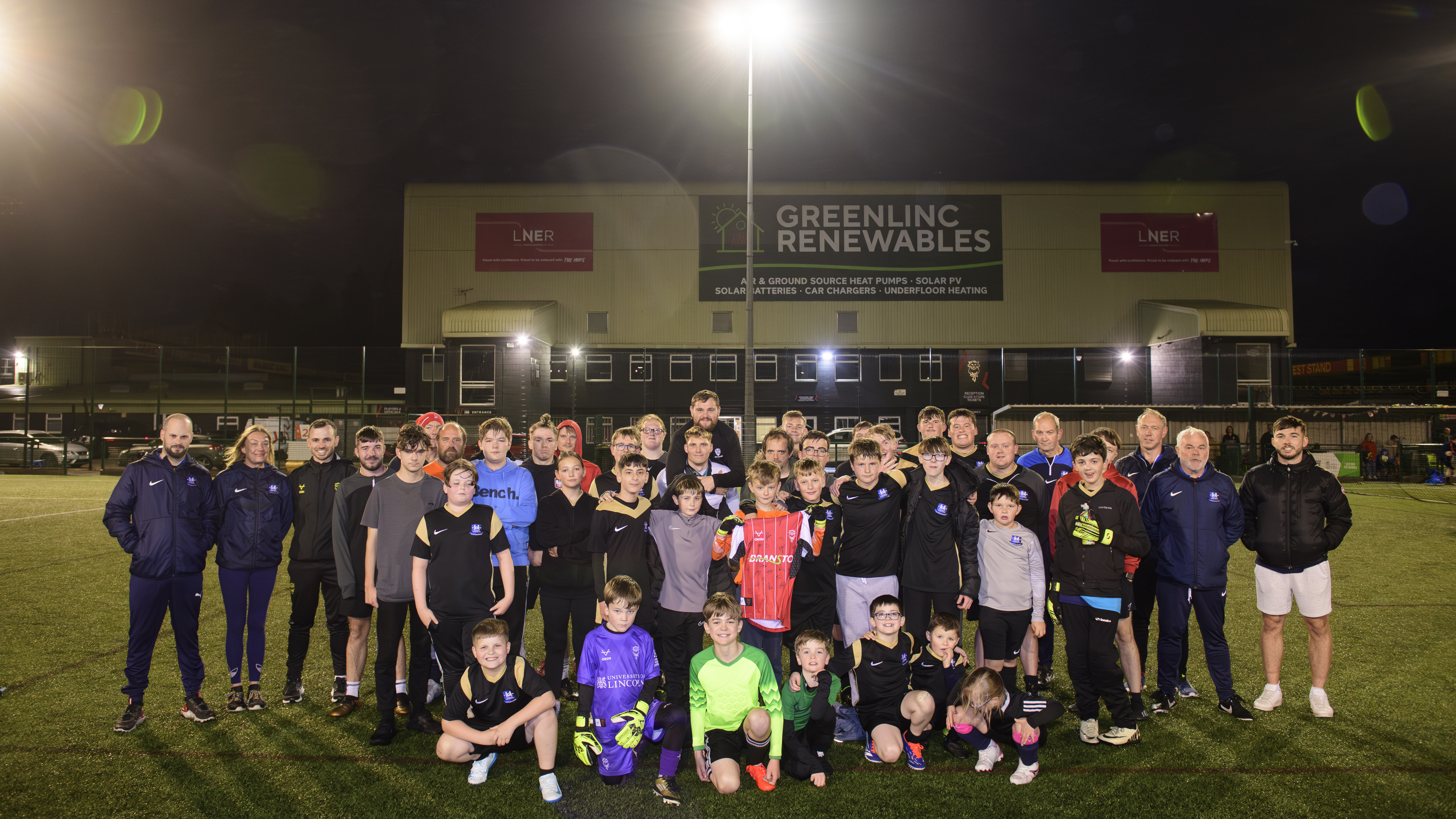 A group of around 20 people pose on a 3G football pitch in front of the LNER Stadium. They are holding a red Lincoln City home shirt.