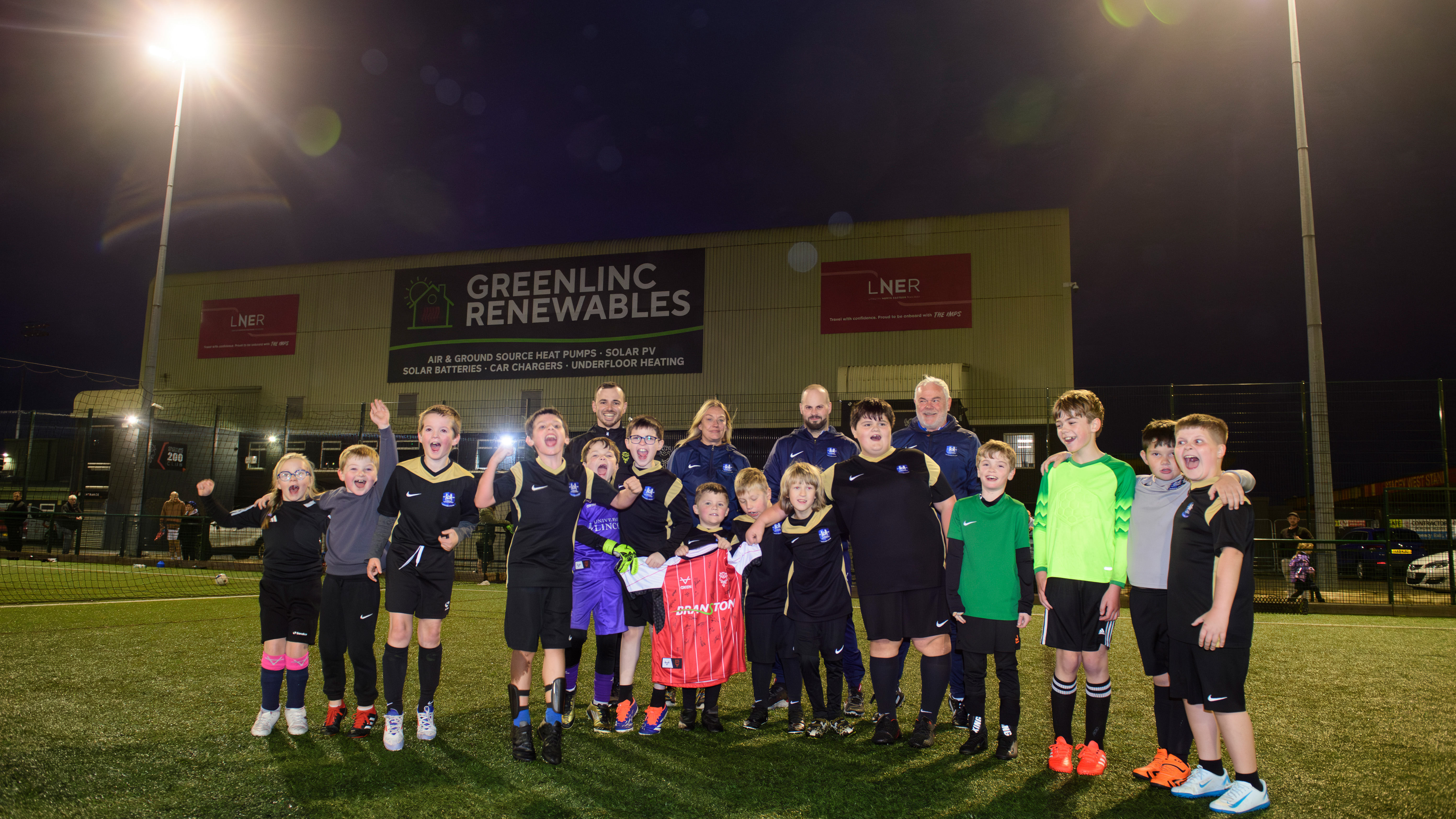 A group of around 20 people pose on a 3G football pitch in front of the LNER Stadium. They are holding a red Lincoln City home shirt.