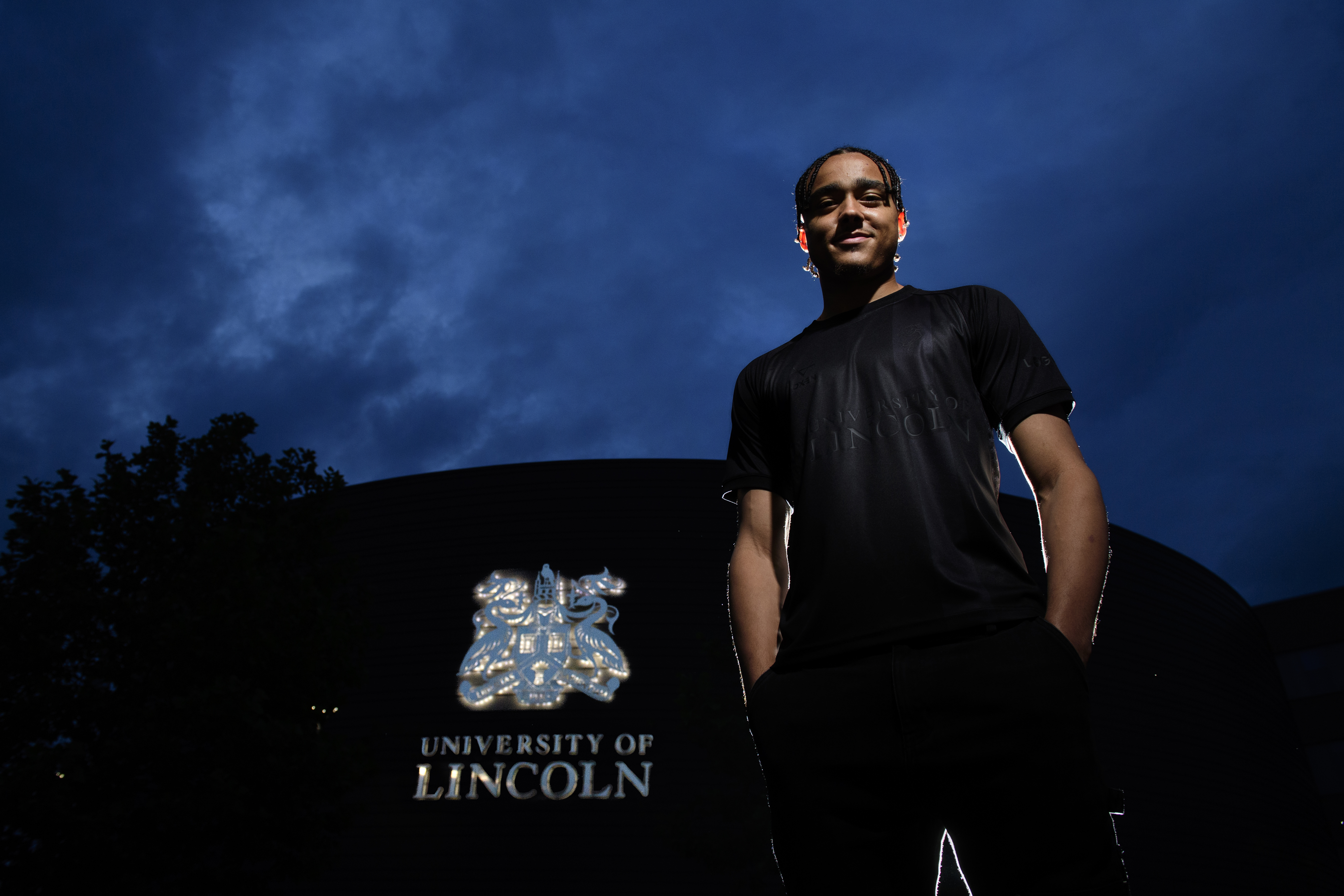 Jovon Makama poses wearing LincolnCIty's all-black third shirt. It is dark and he's stood in front of a building which is all black except for the University of Lincoln logo.