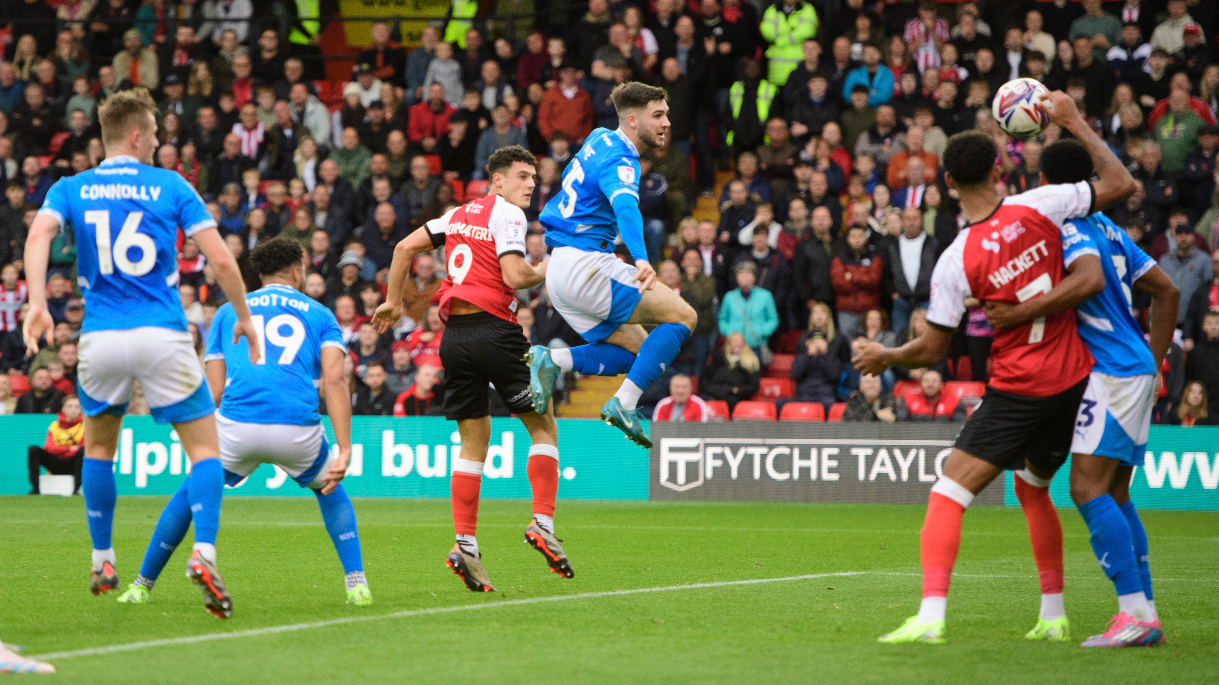 Bailey Cadamarteri heads a goal for Lincoln City. He is mid air, with a Stockport player alongside him, with the ball to his right heading towards the net.