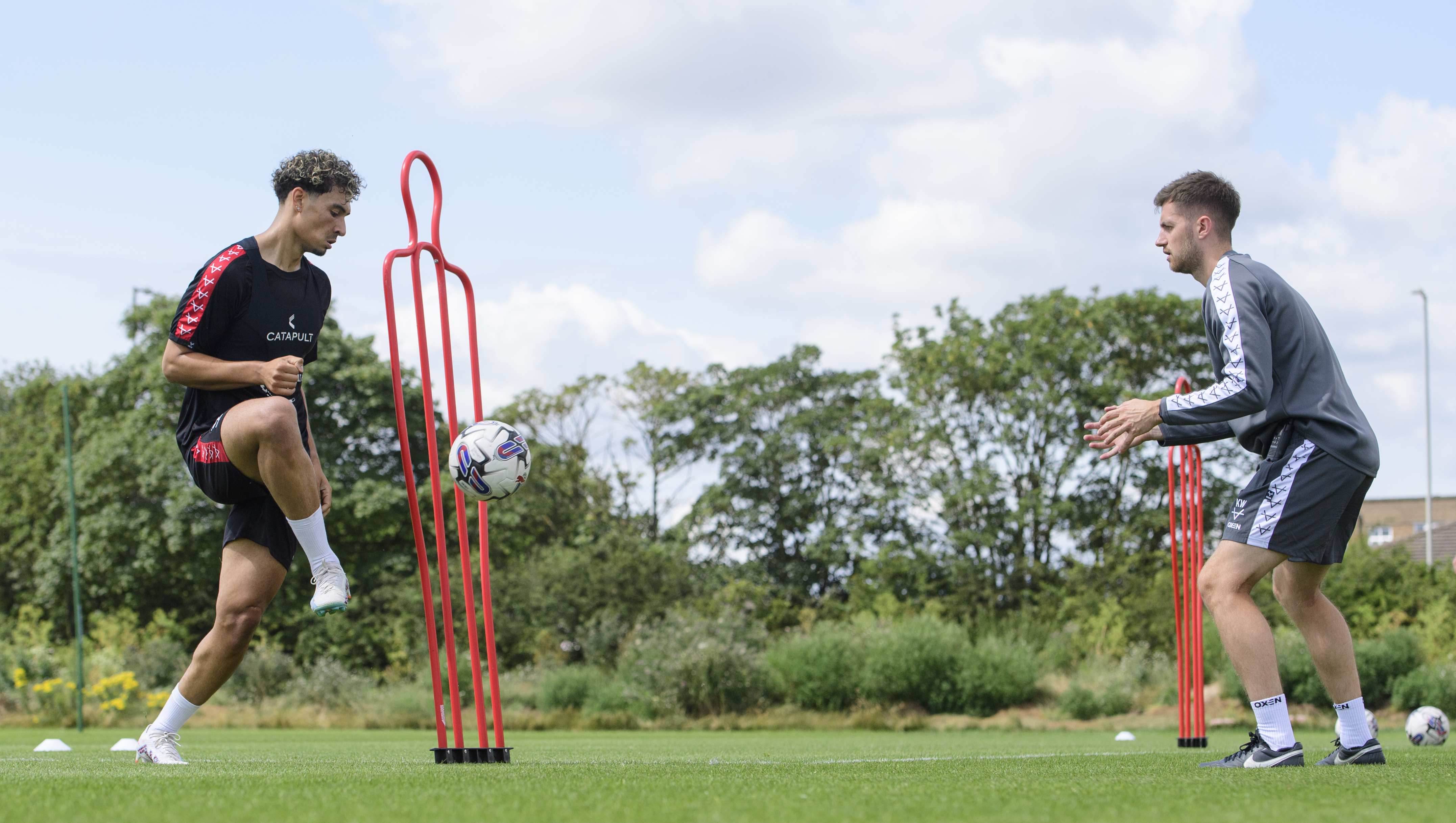 Two men on a football pitch. The man on the left, wearing black training kit, kicks a football back to the man on the right who is wearing a grey training kit.