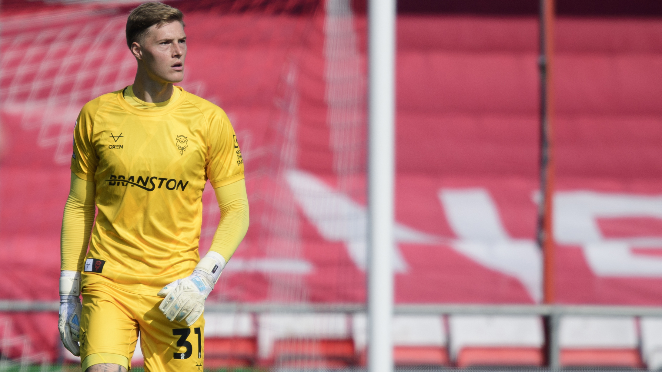 A goalkeeper wearing an all-yellow kit stands watching a game of football. There is a red branded sign in the background.