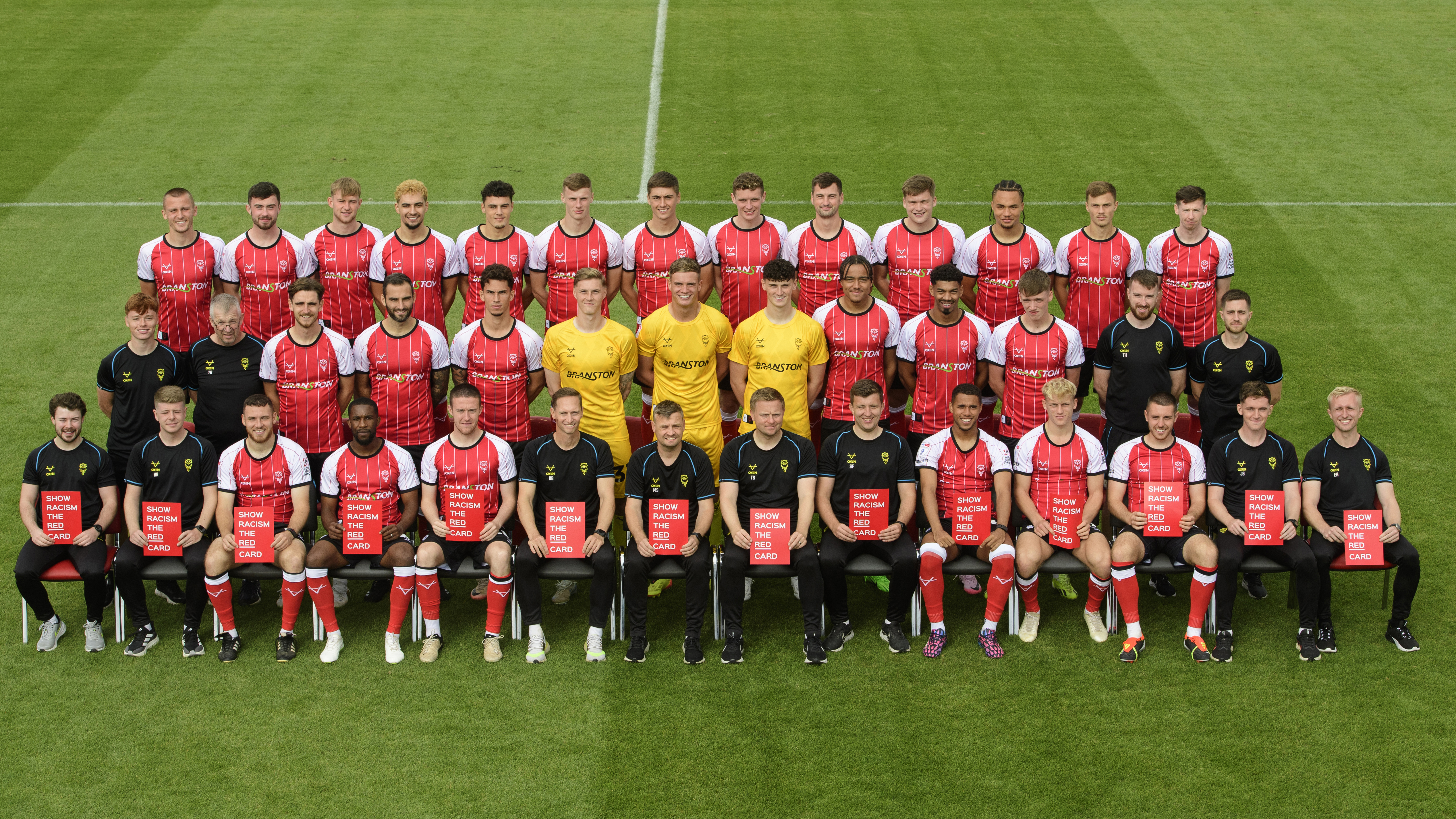 Lincoln City players and coaching staff pose for a team photo. Everyone on the front row is holding up a red sign which reads "Show racism the red card". There are three rows of people, with outfield players in red home shirts, goalkeepers in yellow shirts and staff in black kit.