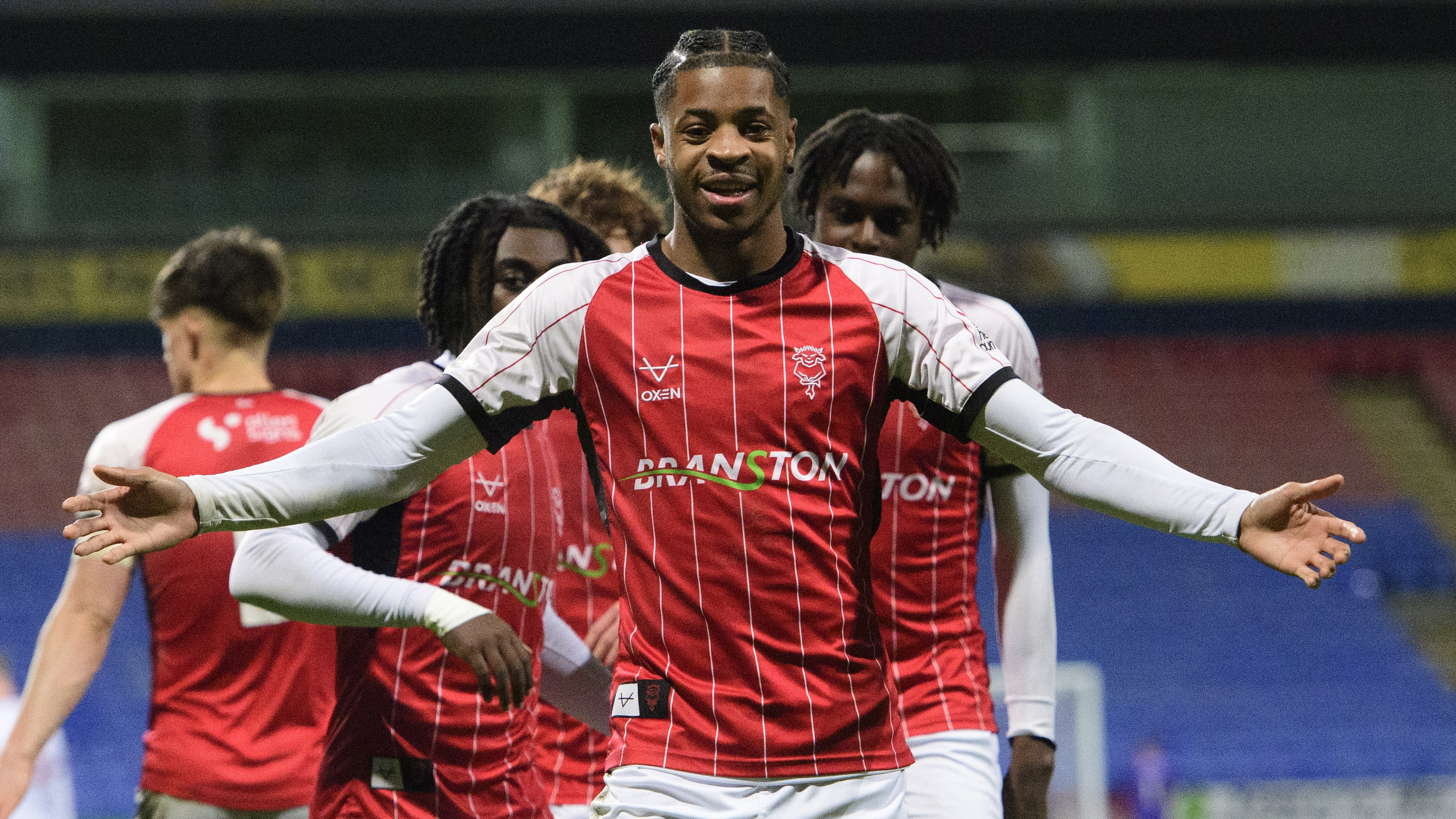 Dakara Wifa celebrates his goal. Wearing a red top with white sleeves, he is looking towards the camera and holding his arms out