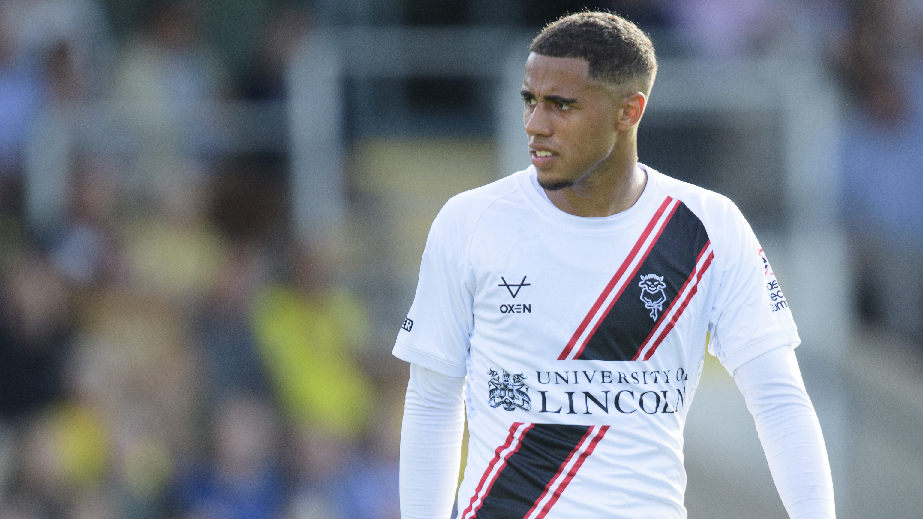 A close up image Ethan Erhahon. He is looking from right to left and wearing a while football top with "University of Lincoln" as the sponsor written on the front.