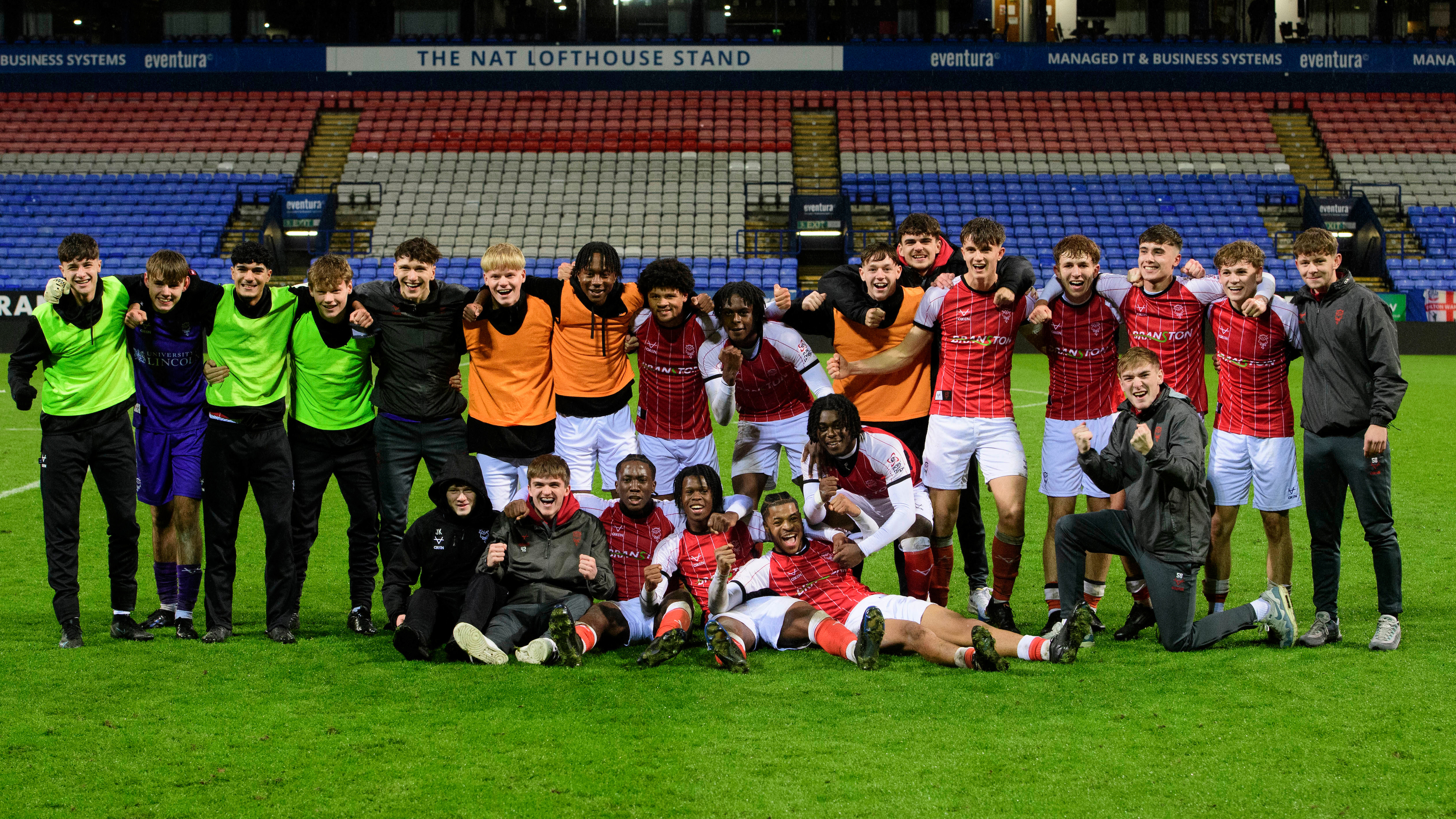 Lincoln City U18 players pose for a group picture after winning at Bolton. They are stood on a green football pitch with seats in the background.