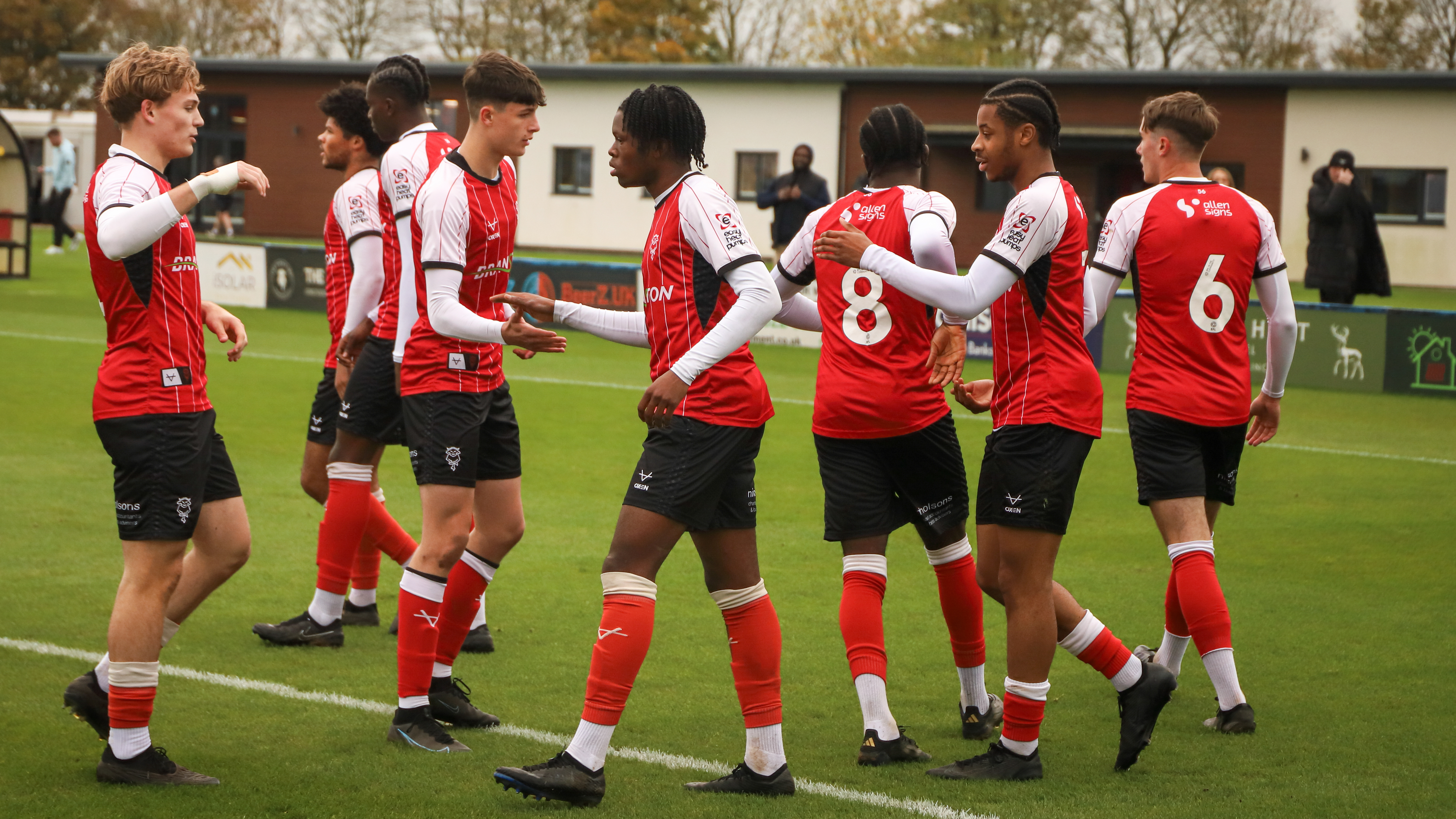 Lincoln City U18 players celebrate a goal. There are eight players, all wearing red shirts and black shorts. They are stood on a grass pitch and in the background is a building.