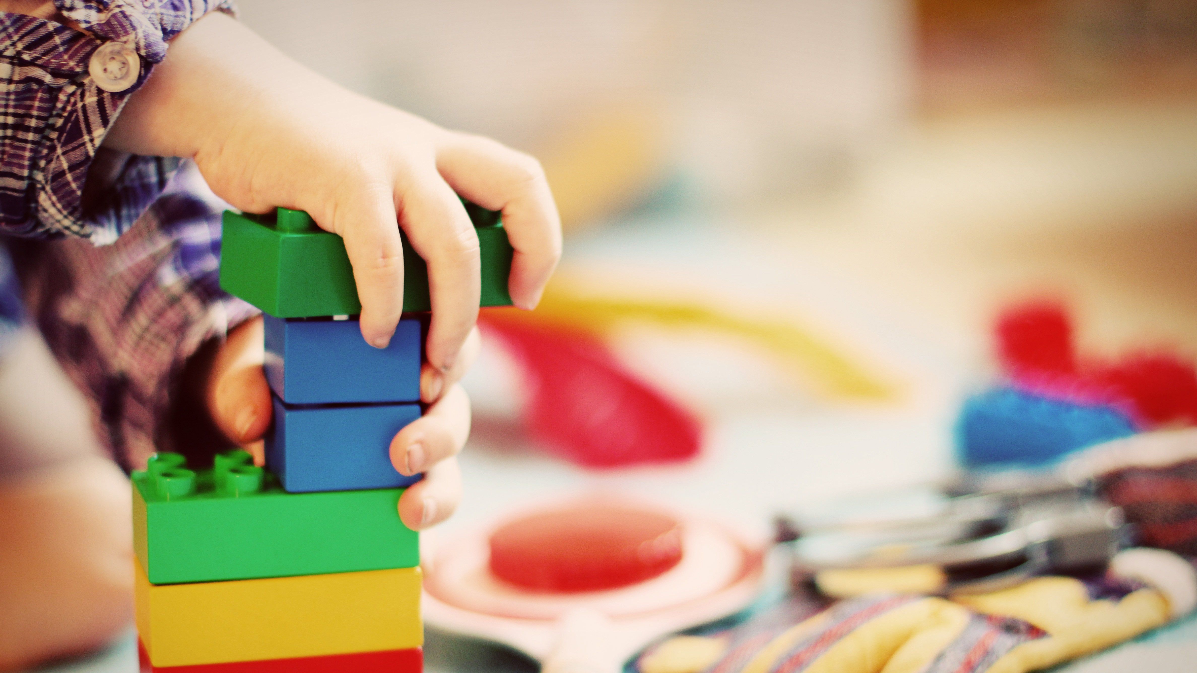 A child's hand plays with building blocks. There are other toys out of focus in the background of the image.