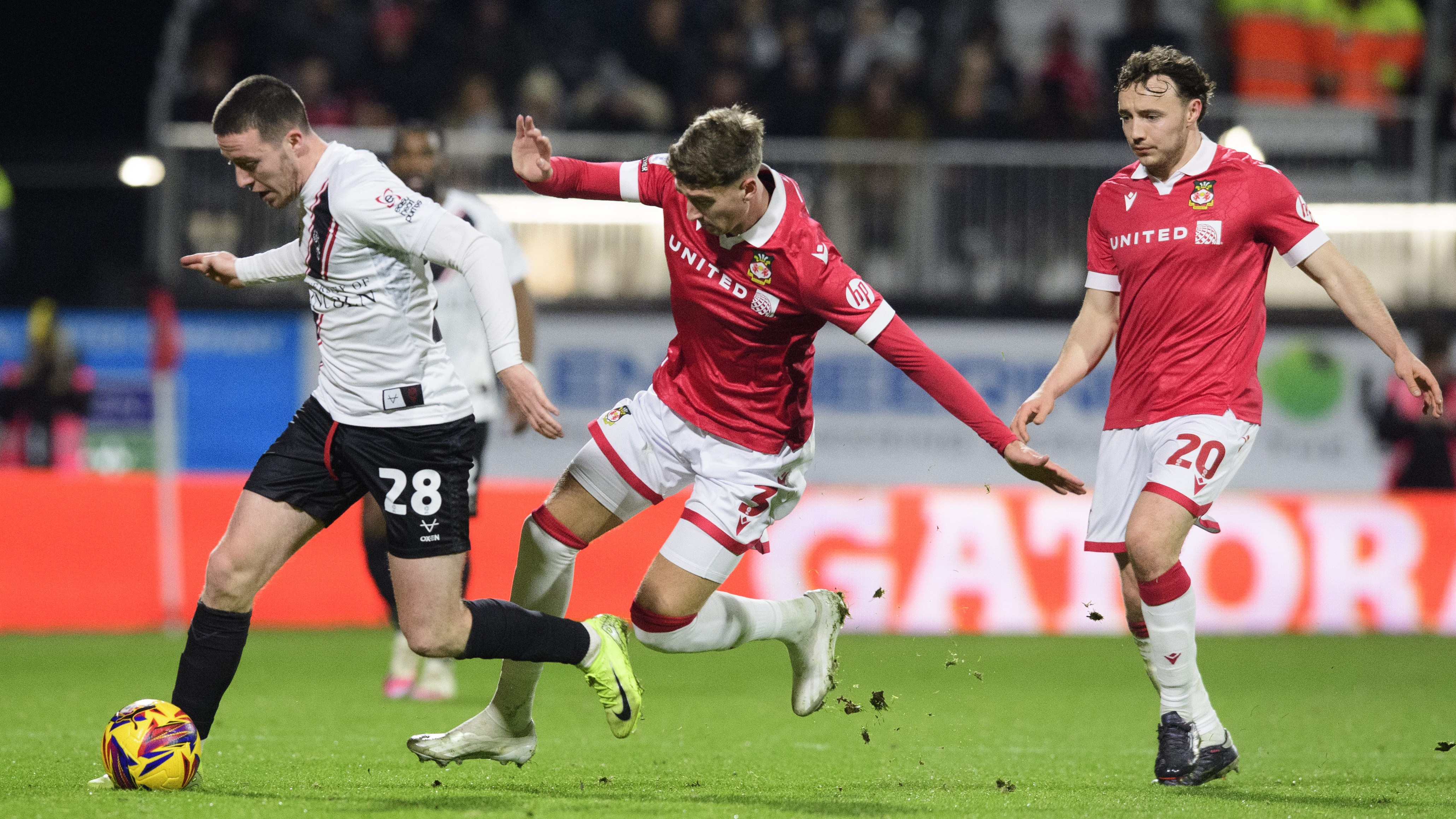 Jack Moylan, wearing a white shirt and black shorts, tries to get away from a Wrexham player wearing a red shirt and white shorts.