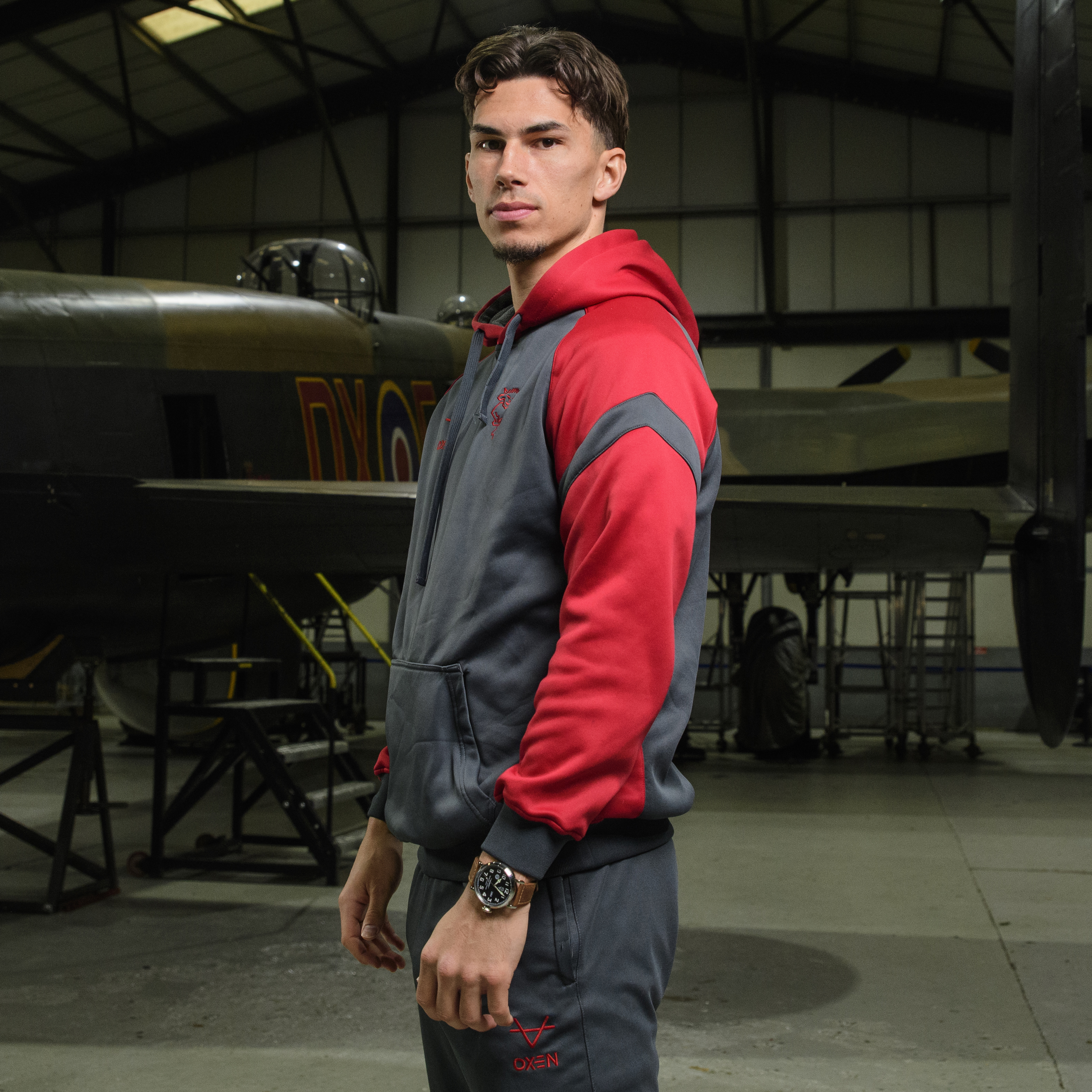 Lewis Montsma stands in front of a plane. He is wearing a tracksuit and wearing a Lincoln City watch