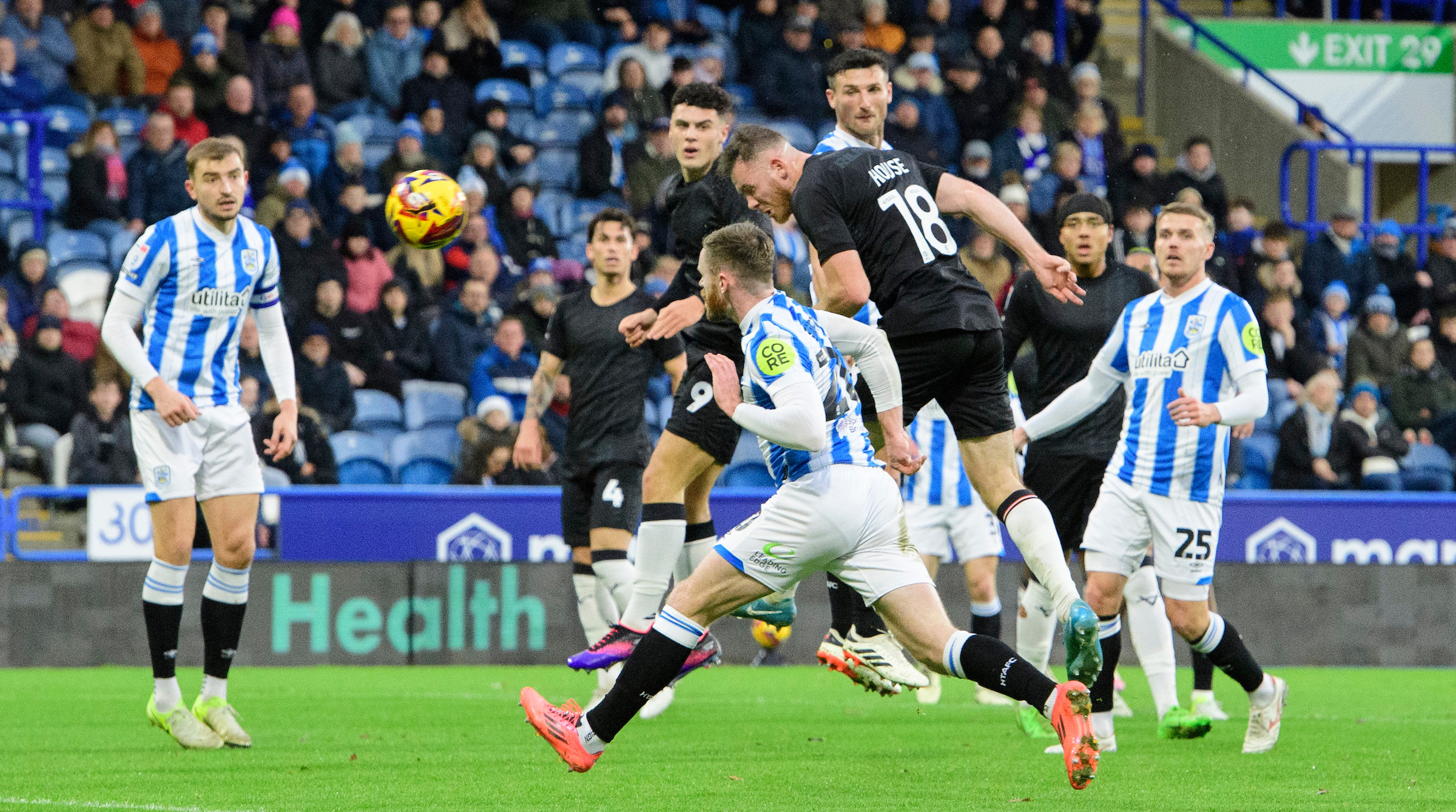 Ben House, wearing an all black kit, jumps highest to head home the opening goal for Lincoln City. In the foreground are several players in red and blue striped shirts.