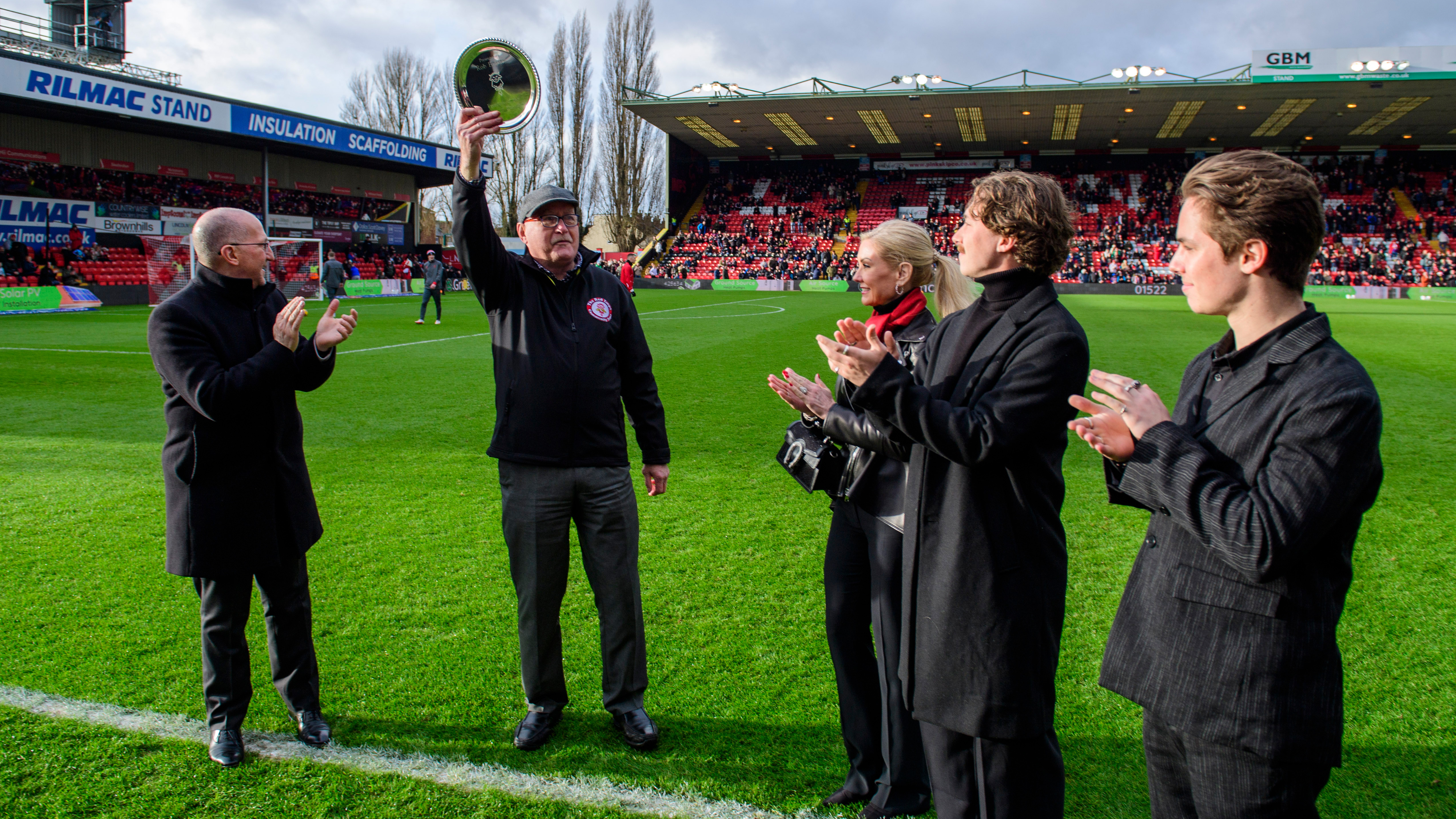 A man holds a silver salver up to salute the crowd while four other people watch and applaiud