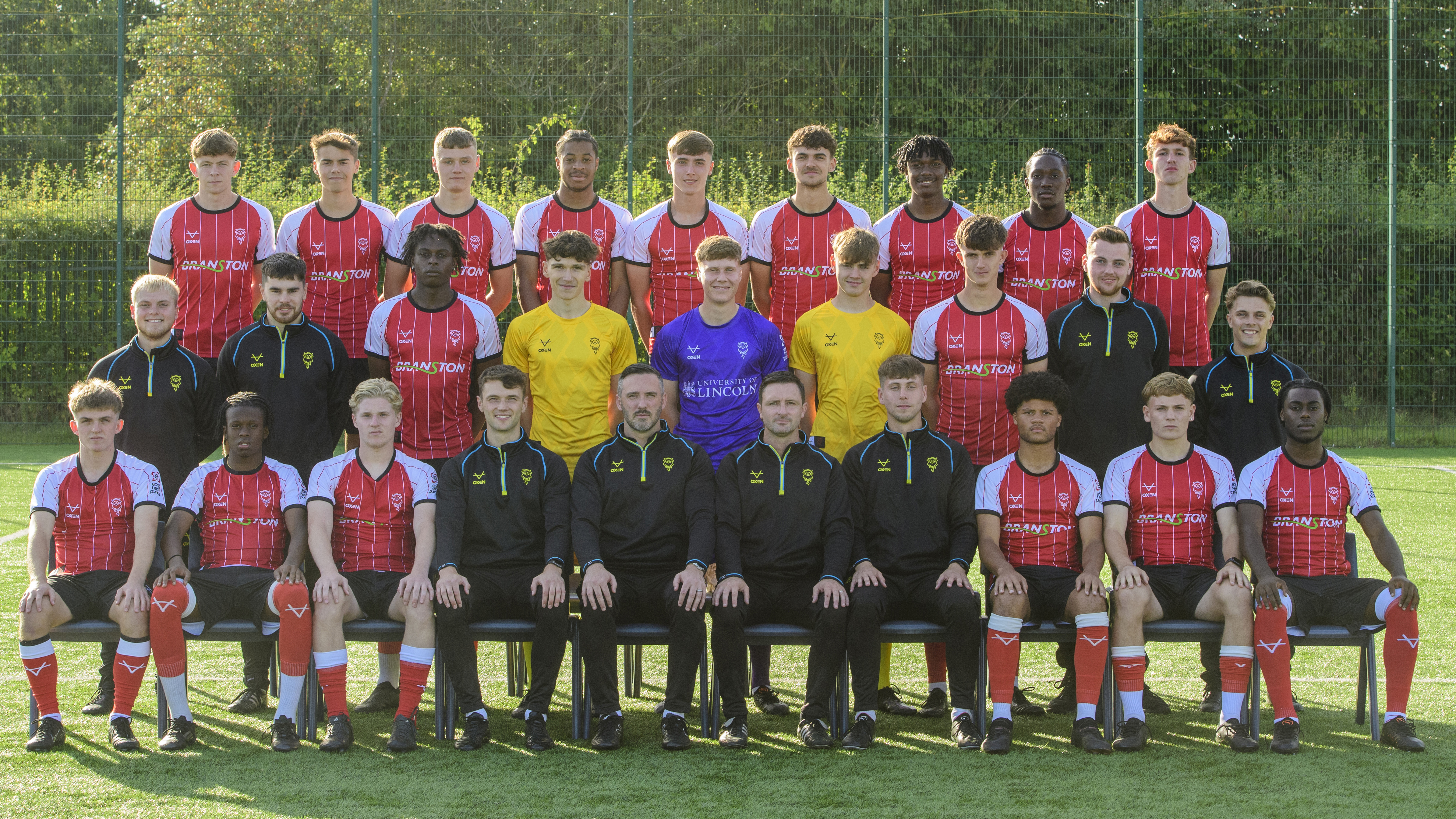 A group photo of Lincoln City's U18 side. Players and staff are stood in three rows facing the camera. Outfield players are in red shirts and black shorts, with the goalkeepers in yellow or purple shirts and staff in all black.