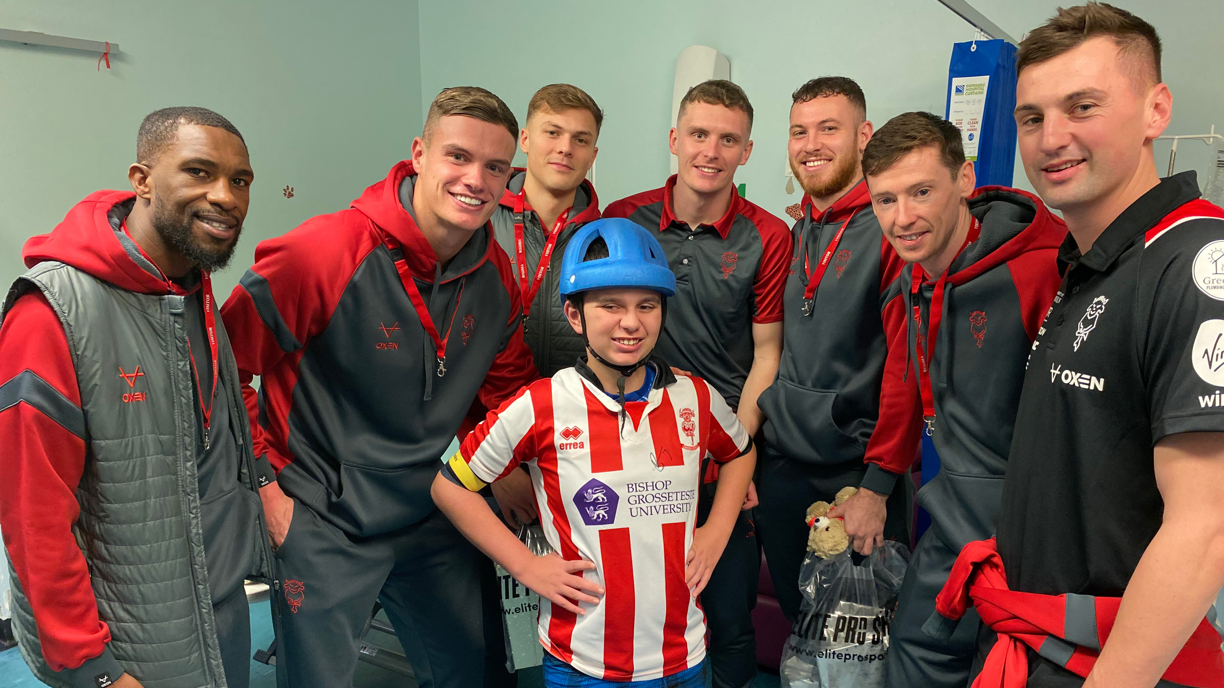 Seven footballers wearing grey and red tracksuits pose for a photo with a boy who is wearing a Lincoln City top.