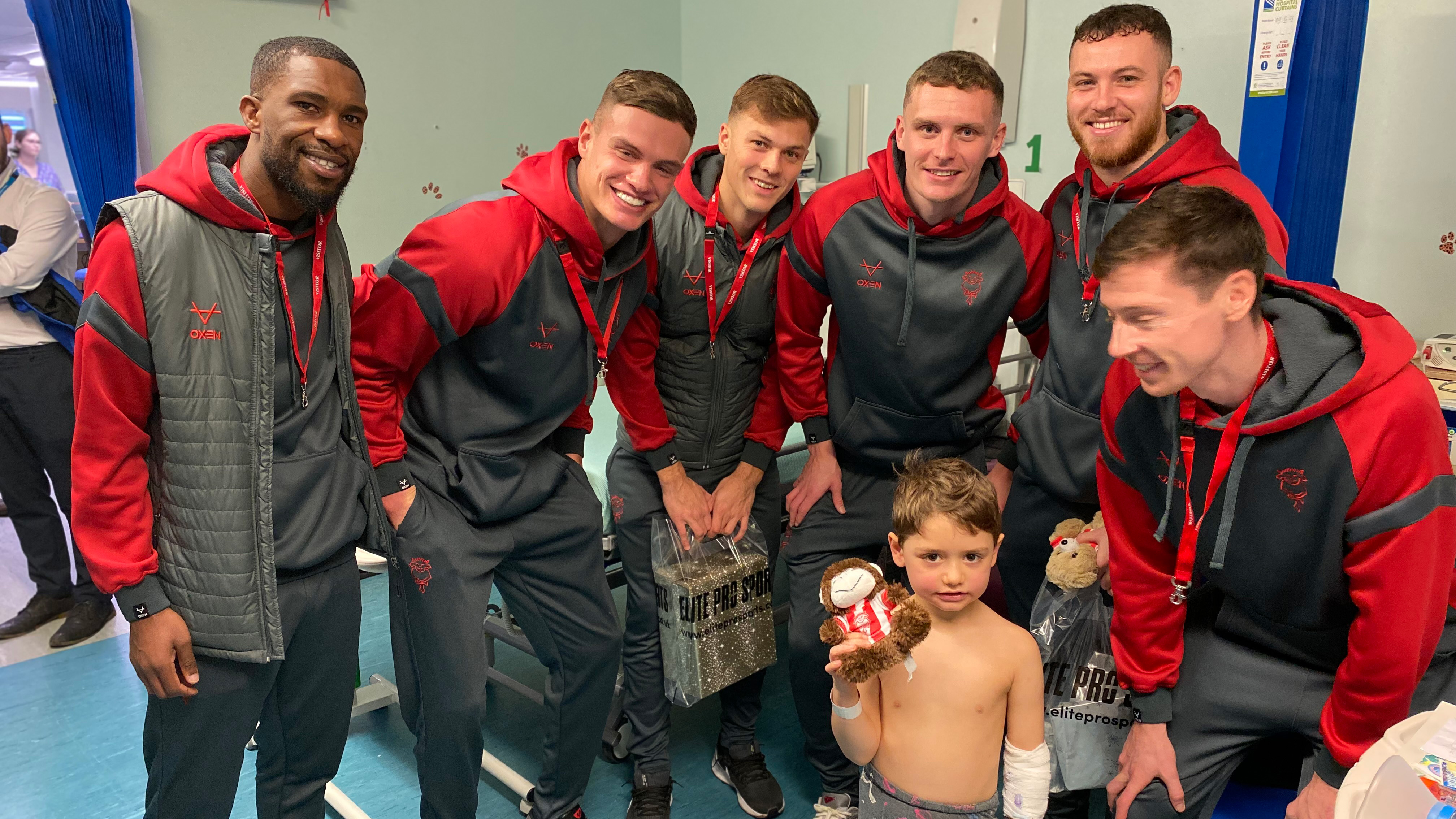 Six men wearing Lincoln City tracksuits pose with a small child who is holding a teddy bear