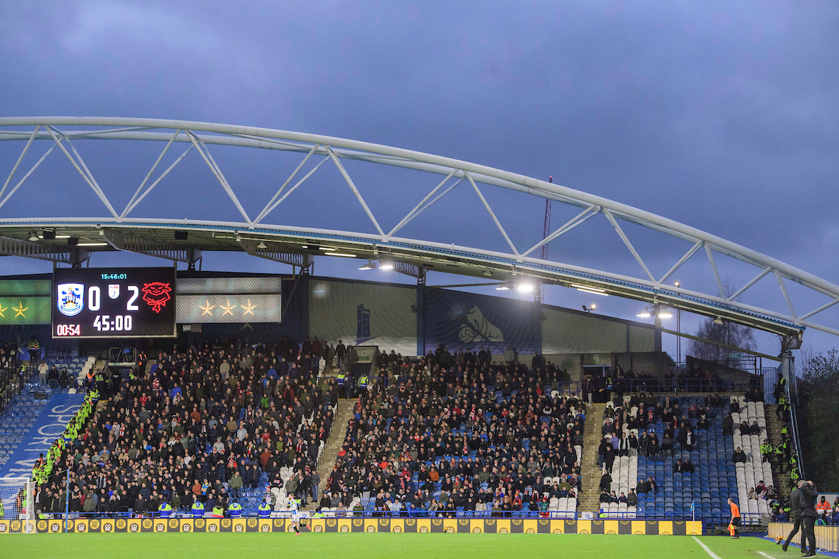 More than 1,300 fans pack out the away end at John Smith's Stadium. In front of them is the pitch and above them is the arch of the sadium roof and a cloudy sky. 