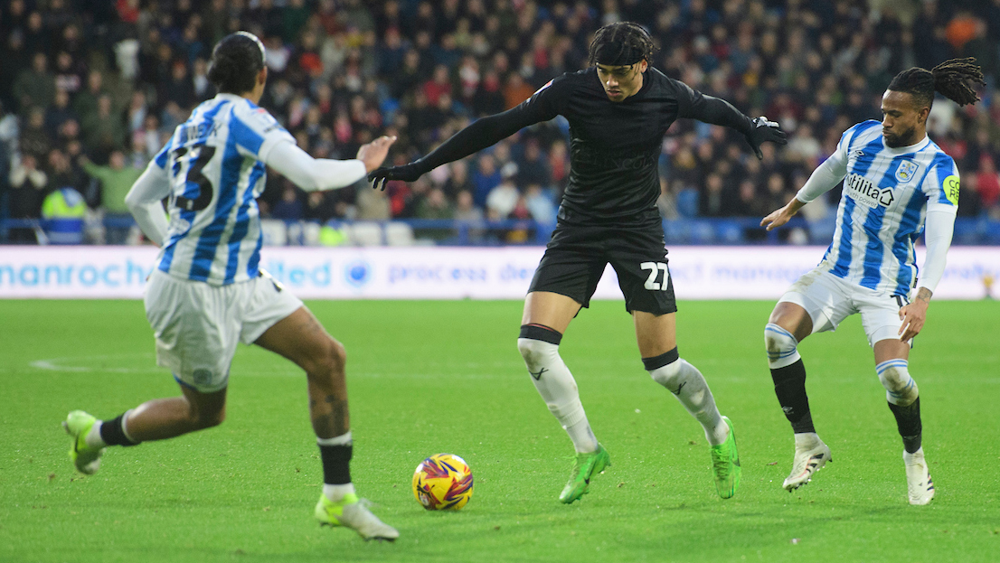 Jovon Makama, in an all black kit, dribbles with a football with Huddersfield players in blue and white shirts on either side of him.