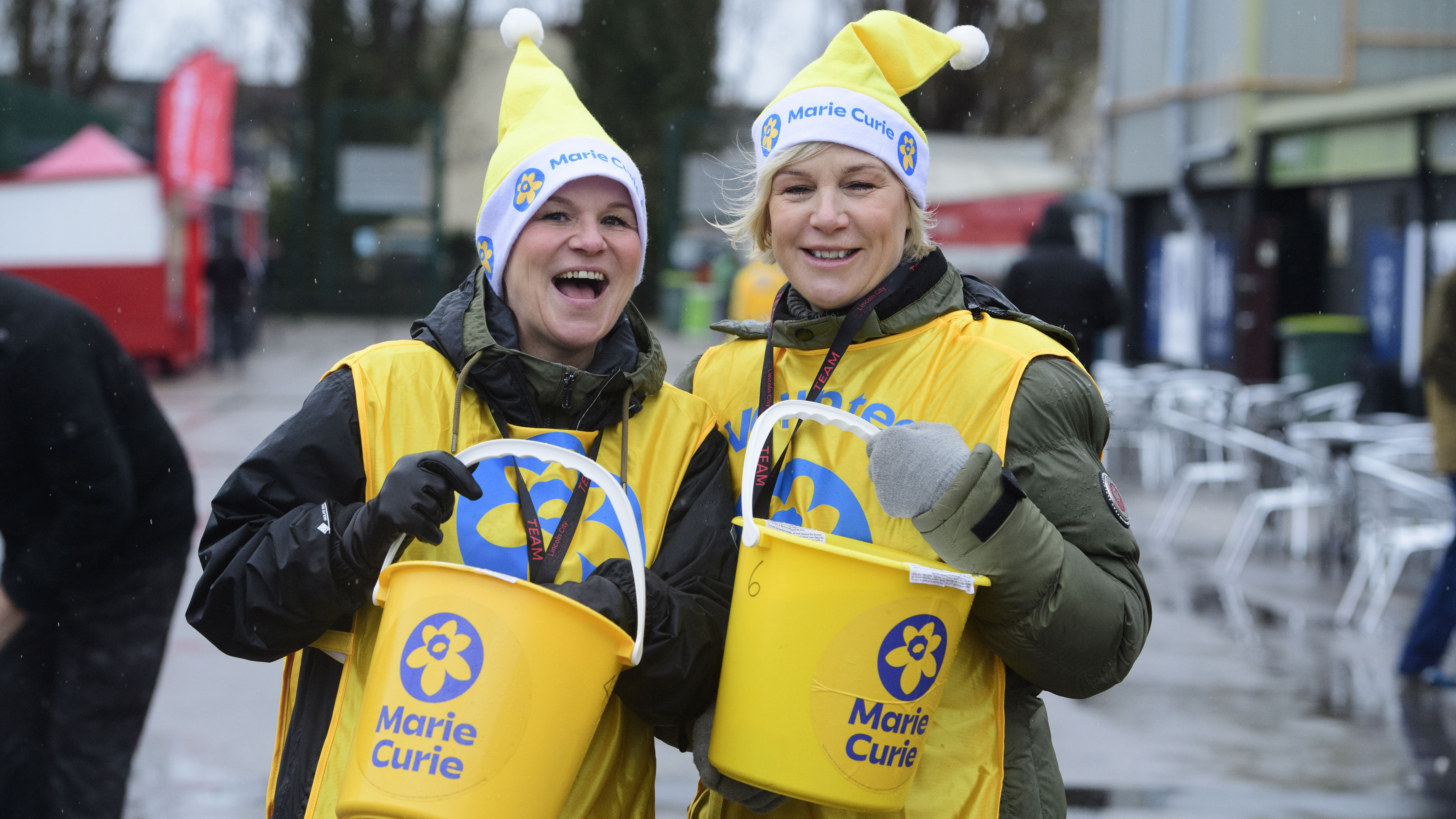 Two women dressed in yellow hold yellow buckets with "Marie Curie" written on them.