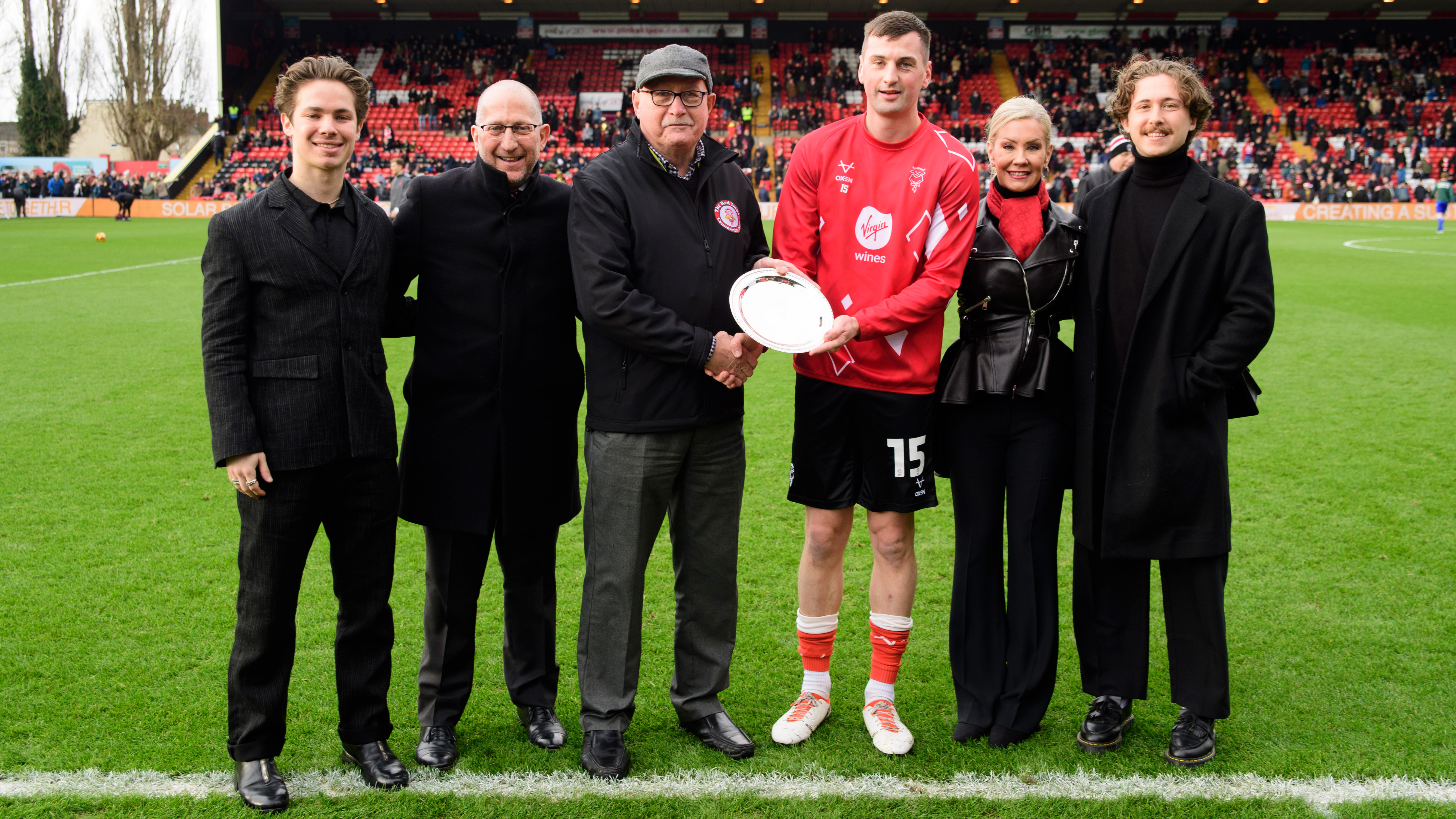 A group of people stand beside a football pitch. Stood in the middle is a man who is being presented with a silver salver.