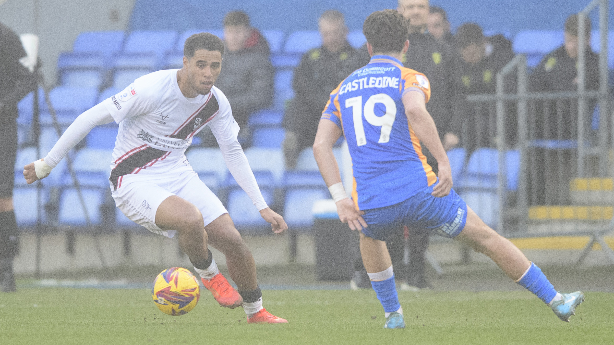 Ethan Erhahon, wearing a predominantly white kit, runs with the ball. In front of him is a Shrewsbury TOwn player in a predominantly blue kit with yellow sleeves.