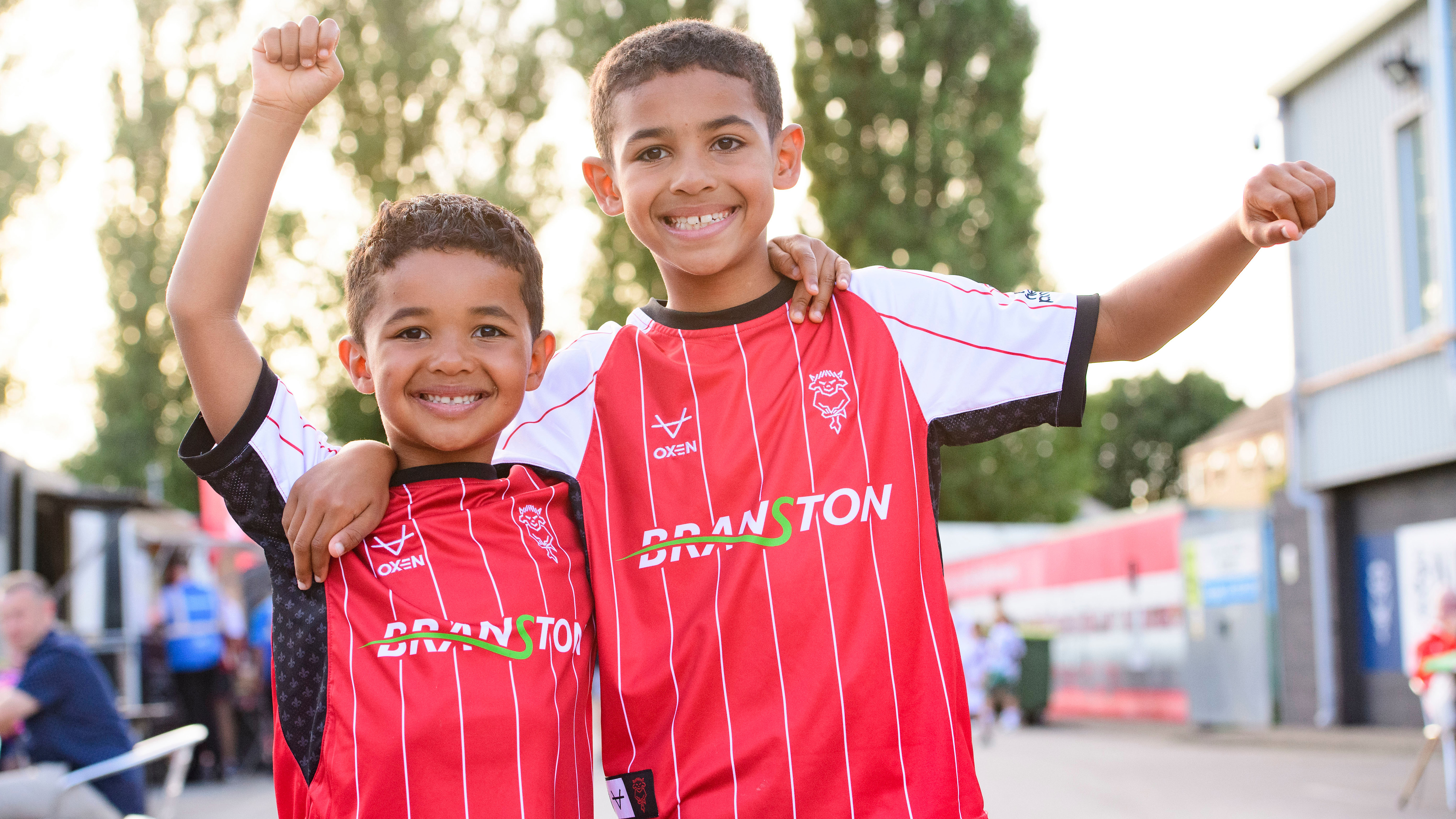 Two young boys pose for the camera. They are wearing red replica Lincoln City tops.