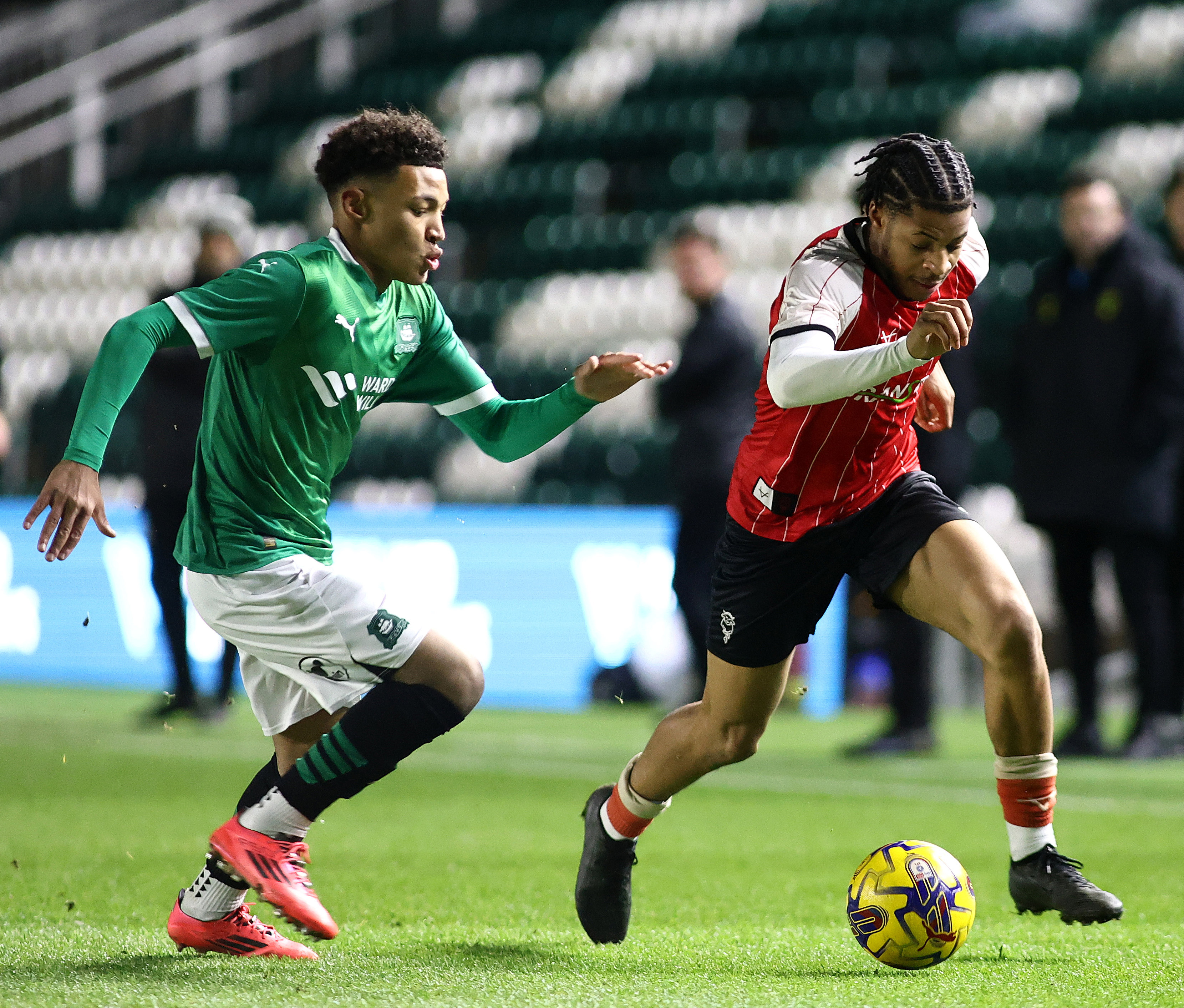 A footballer in a red shirt and black shorts tries to get away from a footballer wearing a green shirt and white shorts. There is a ball in front of the player on the right in the red shirt.