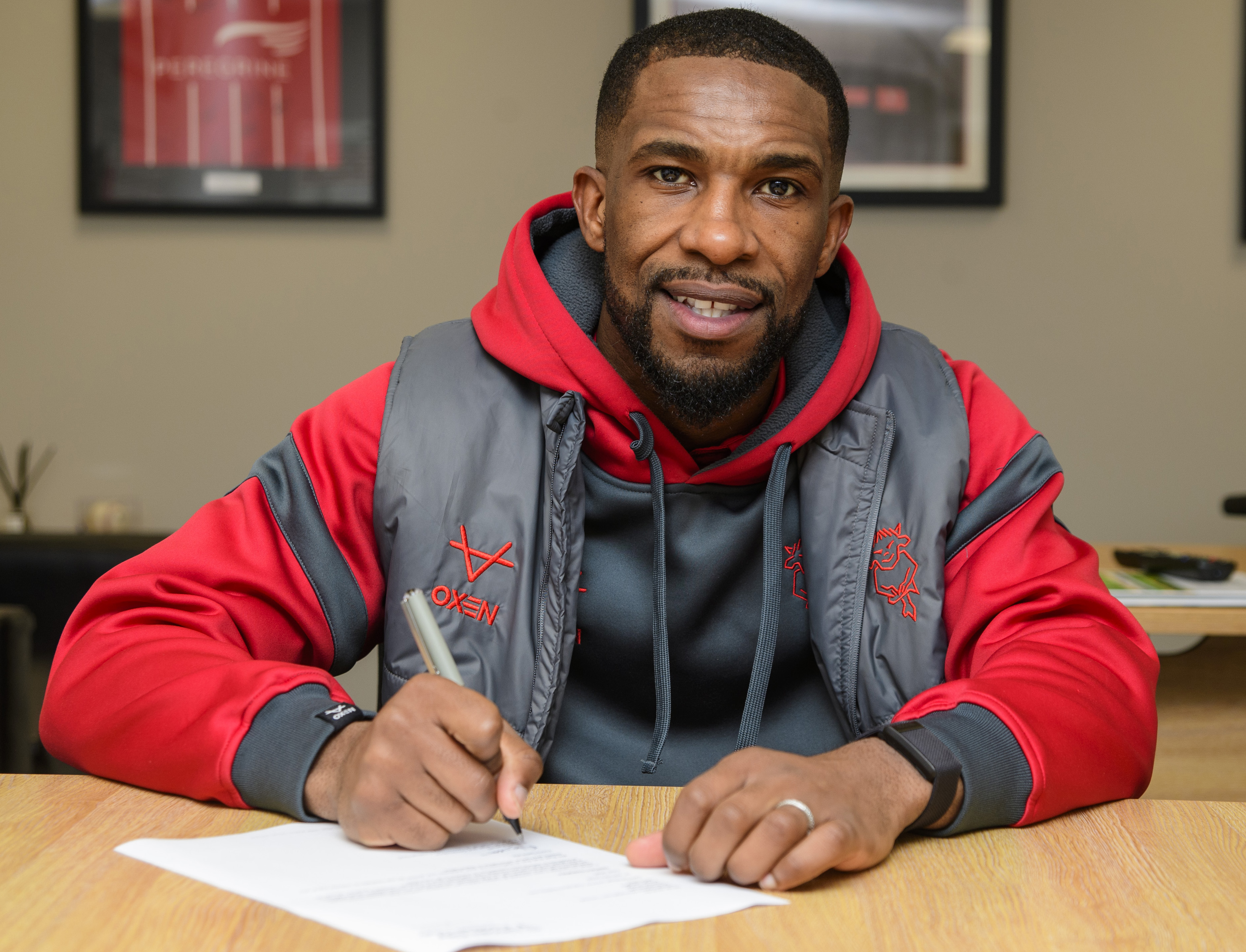 Tendayi Darikwa sits at a desk to sign a new contract. He is holding a pen and wearing a grey and red hoodie while looking at the camera..