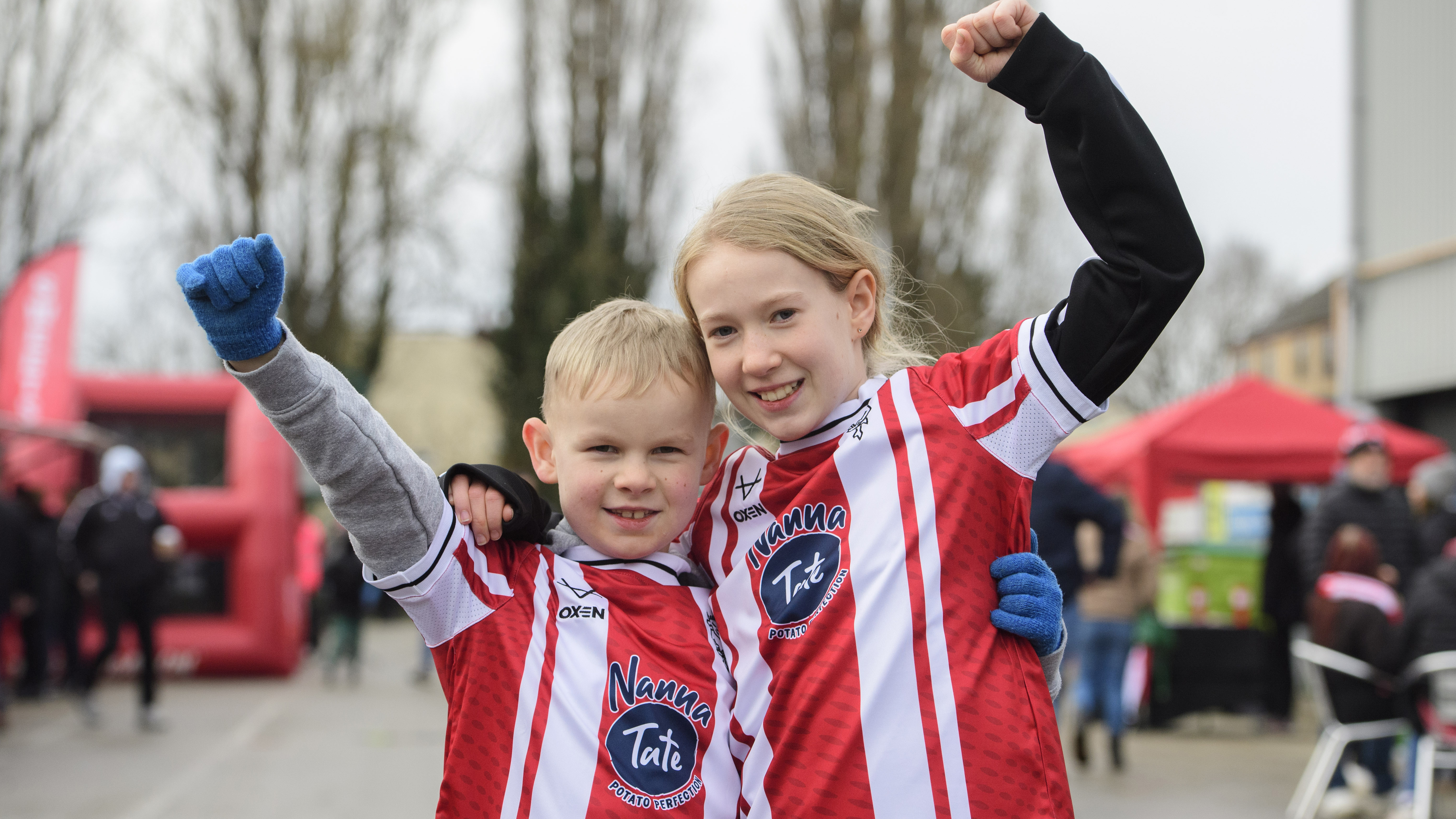 Two children pose wearing red and white football tops. They are smiling and each holding one arm up and their other arm around the other child.