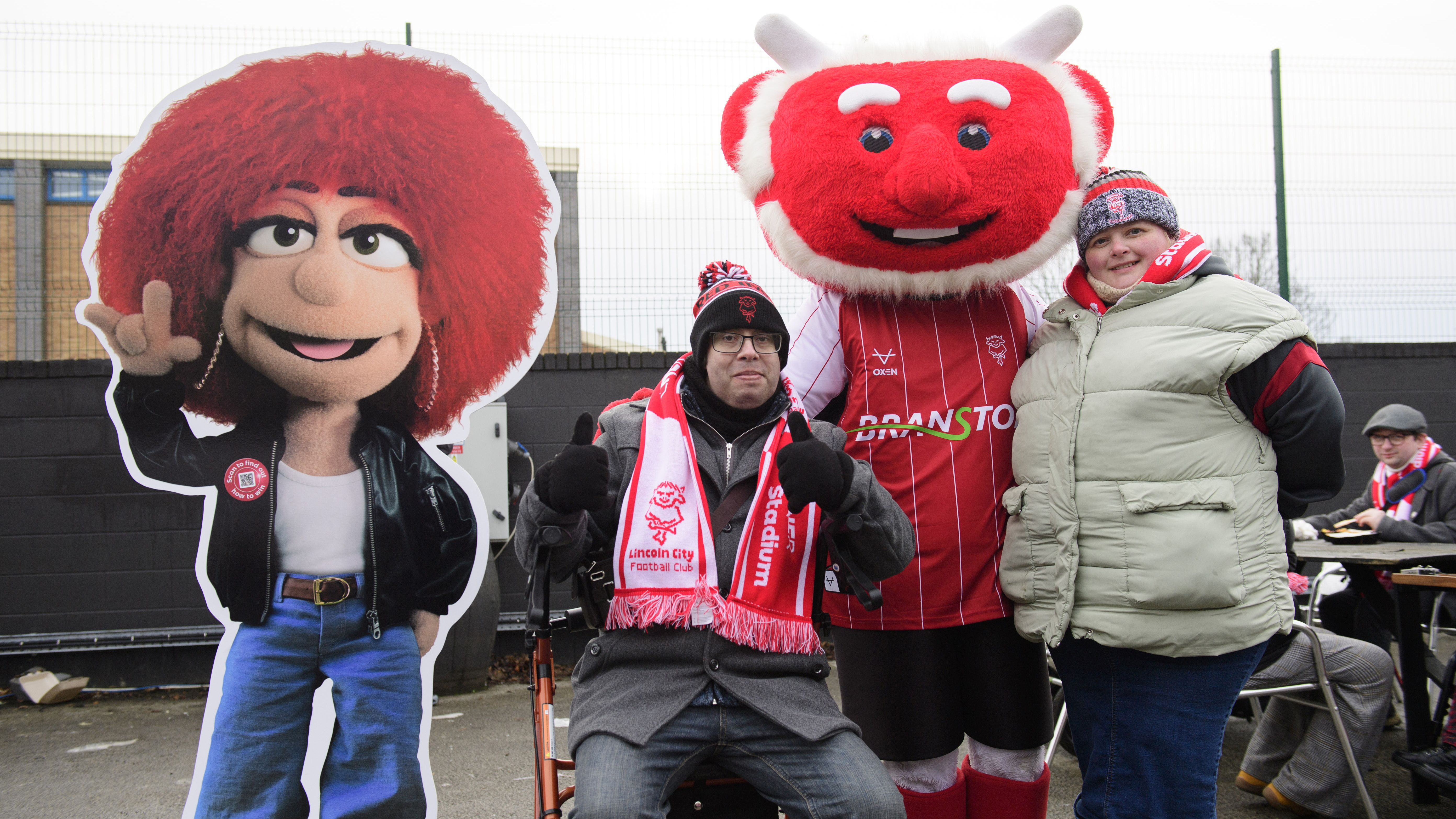 Two Lincoln City supporters pose with Poacher the Imp and with a cutout of Eleanor the LNER mascot