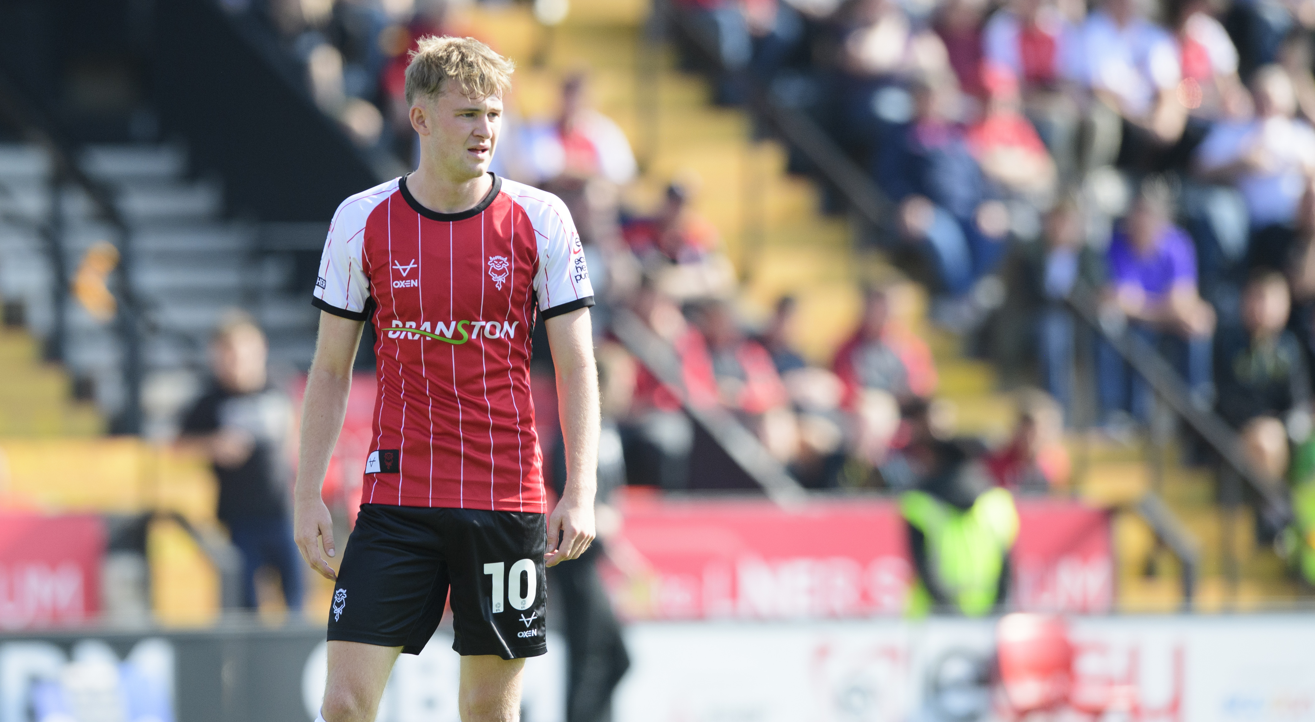 JJ McKiernan stands on a football pitch looking from the left of the image to the right. He is wearing a red football top with "Branston" on the front. In the background is a stand with people watching the match.