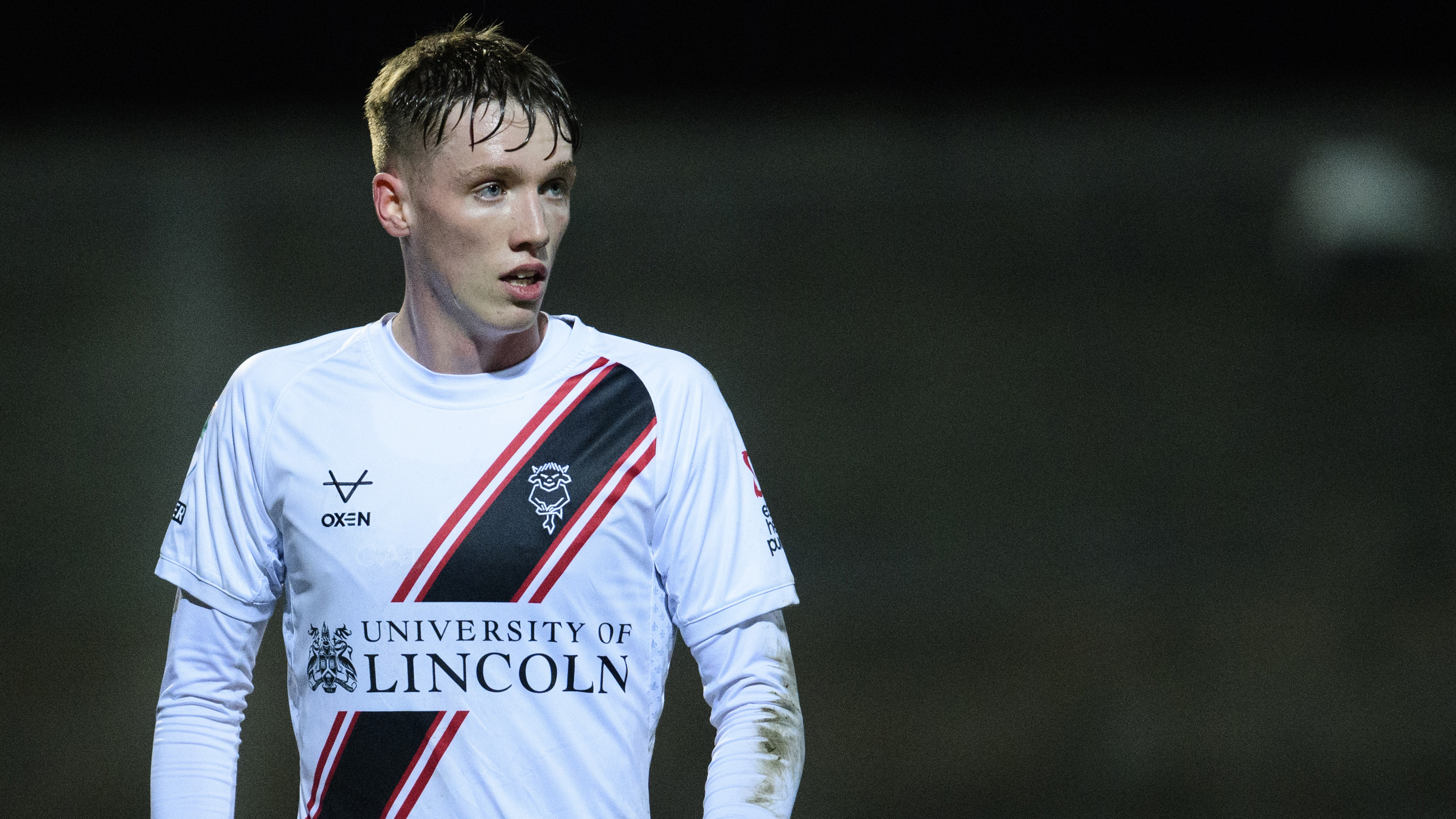 Oisin Gallagher looks from the left of the screen. He is wearing a mostly white kit with "University of Lincoln" on the front.