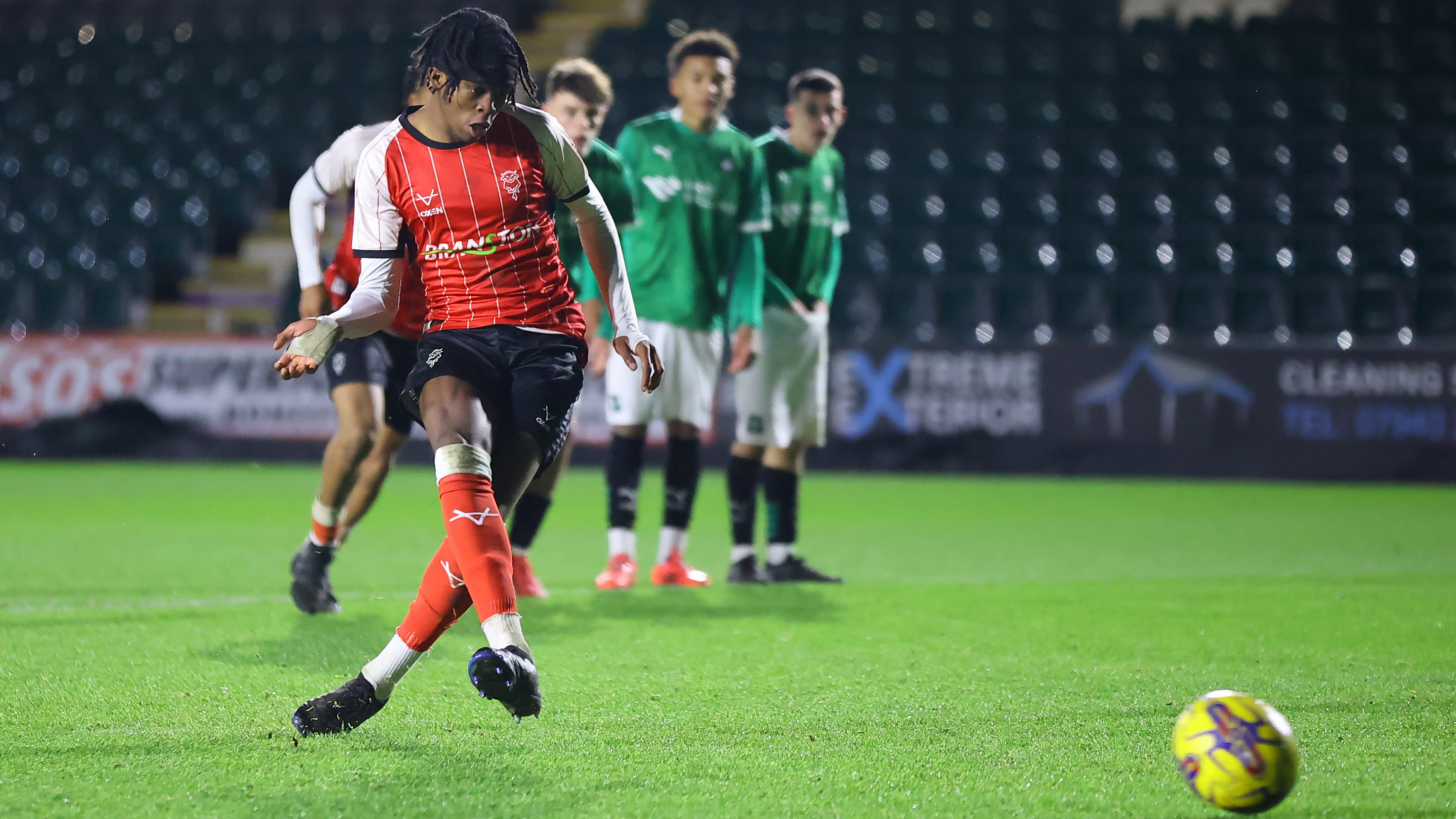 Zane Okoro scores a penalty for Lincoln City U18. He is wearing a red kit and on the left of the picture. He has kicked the ball which is on the bottom right of the image.